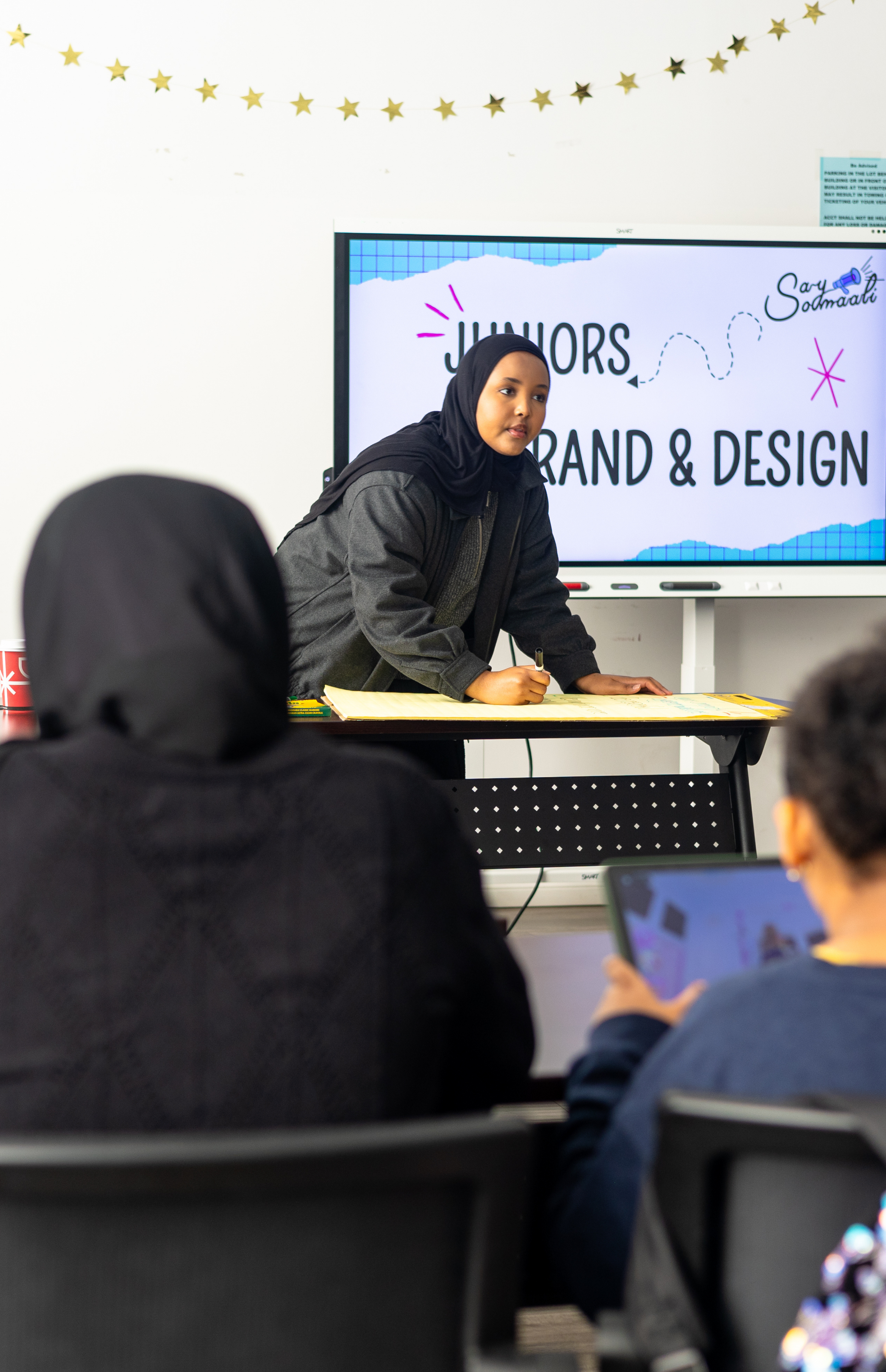 A woman in a black hijab and gray jacket giving a presentation on brand and design to an audience, with a digital screen behind her displaying the words 'Juhiors Brand & Design' and the name Sar Saudmatti.