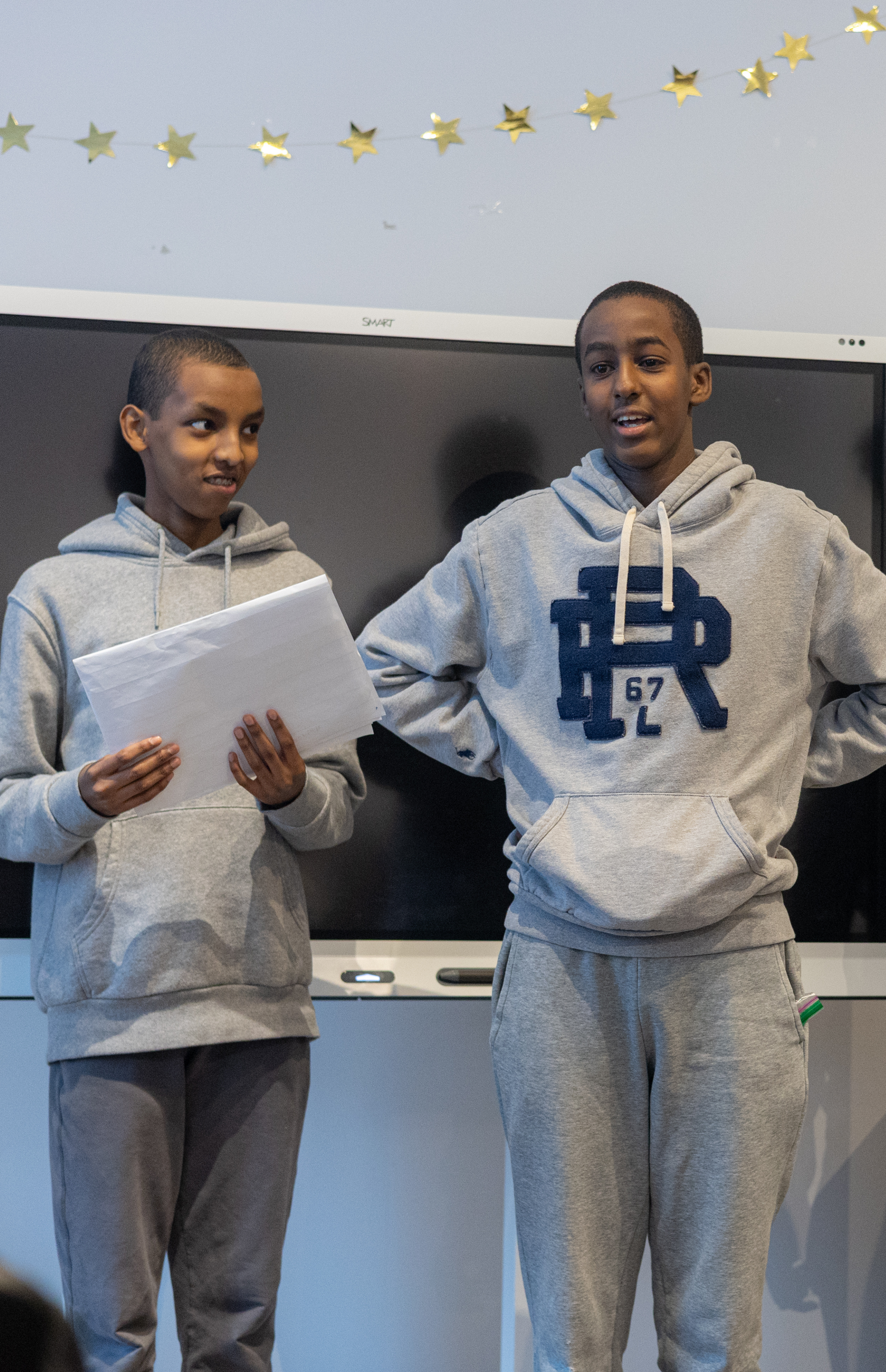 Two boys wearing gray hoodies and sweatpants standing in front of a large screen, one holding papers.