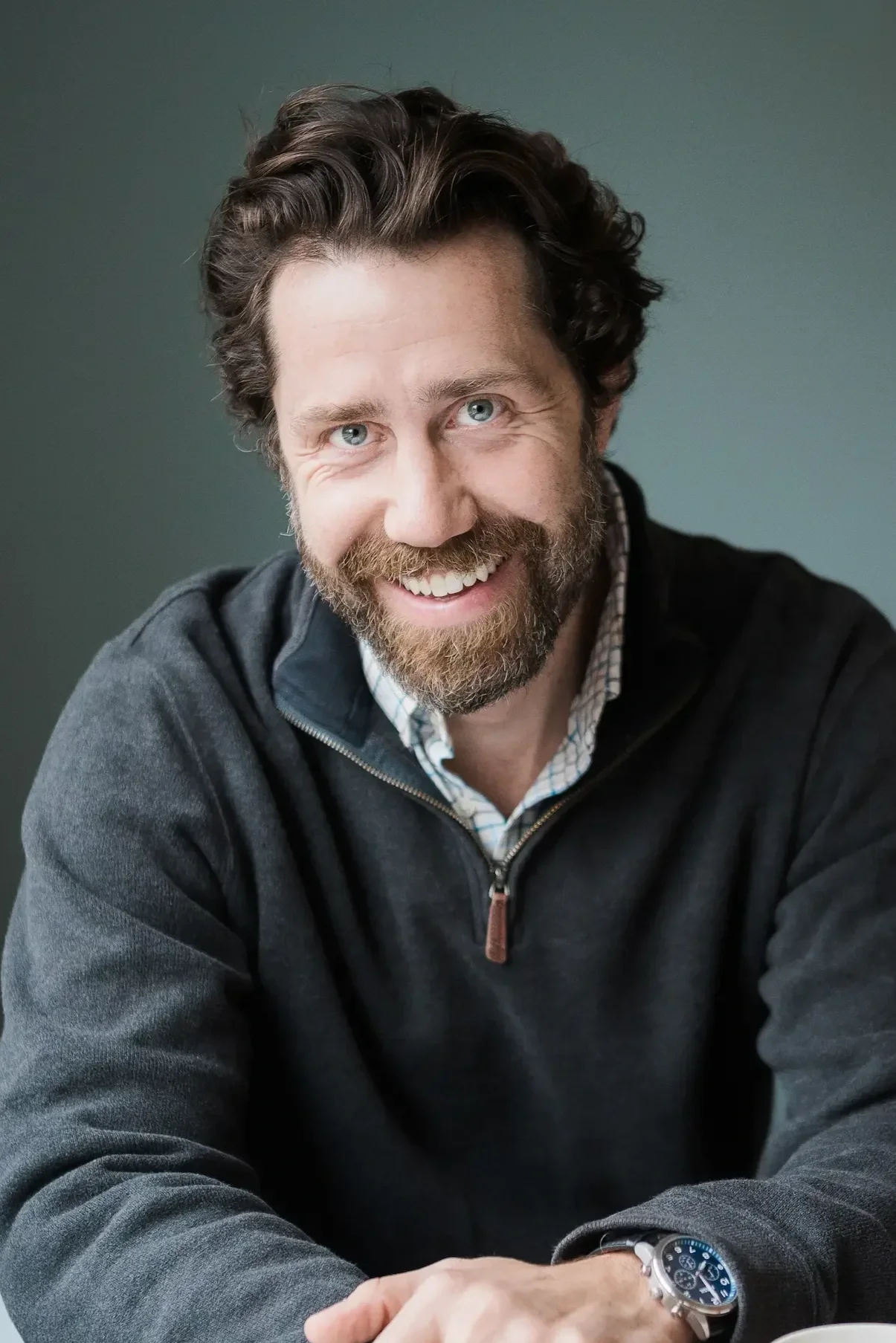 A smiling man with a beard and mustache, wearing a dark zip-up sweater over a checkered shirt, sitting at a table with a gray background.