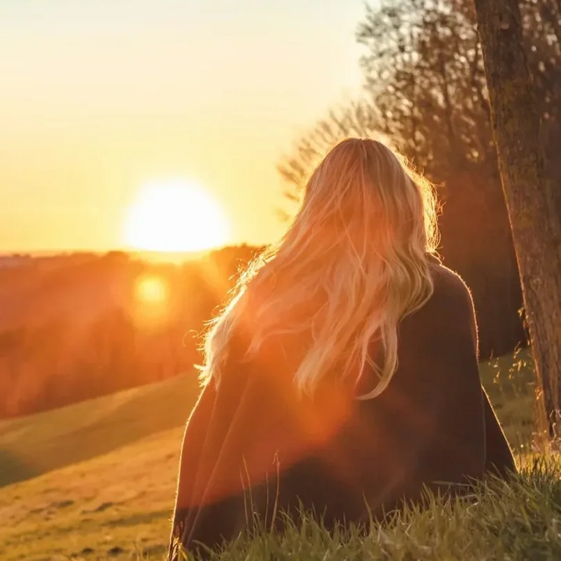 A woman with long blonde hair sitting on grass at sunset, looking away from the camera near a tree.