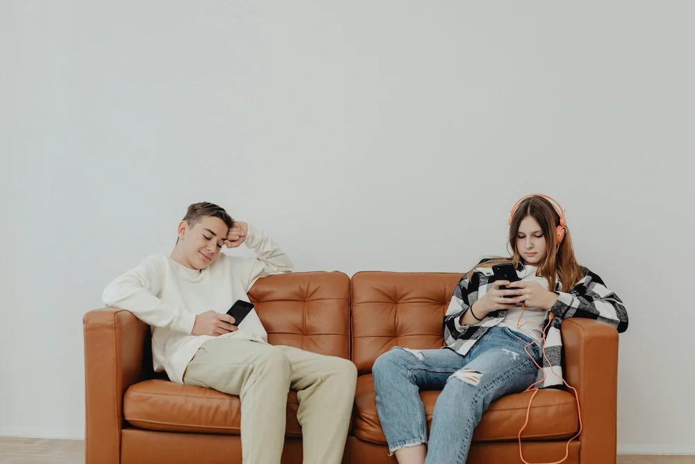 A young man and woman sitting on a brown leather couch, both using smartphones. The woman is wearing headphones, a black and white checkered shirt, and ripped jeans, while the man is wearing a white sweatshirt and beige pants.