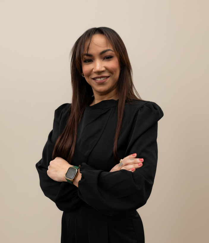 A woman with pink braided hair, wearing large turquoise glasses, a black uniform top, and layered necklaces, standing with arms crossed in front of a white wall.