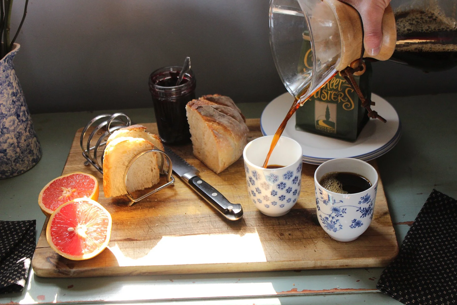 Breakfast setup with sliced bread on a wood cutting board, grapefruit halves, a jar of jam, two cups of black coffee, a knife, a hand pouring coffee from a Chemex, a plate with a coffee bag, and a flowering plant in a blue-spotted pot.
