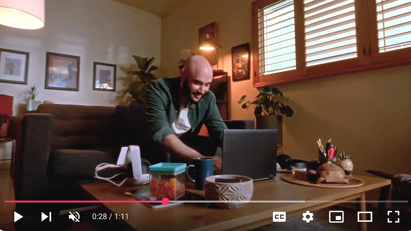 A man smiling and working on a laptop at a coffee table in a cozy living room with plants and framed artwork, with sunlight coming through wooden blinds.