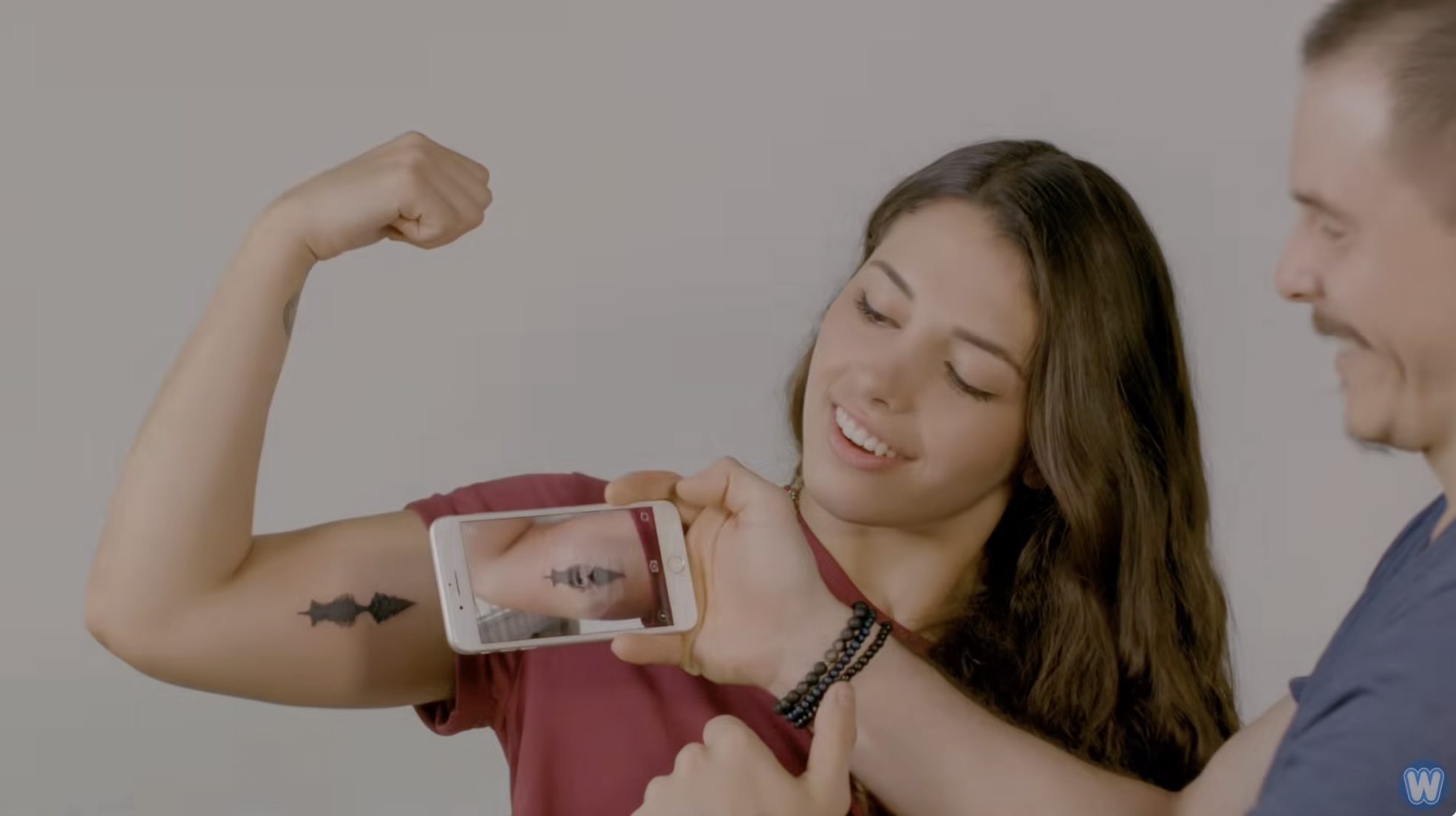 Woman holds her bicep up and partner uses phone to read her tattoo and play a soundwave