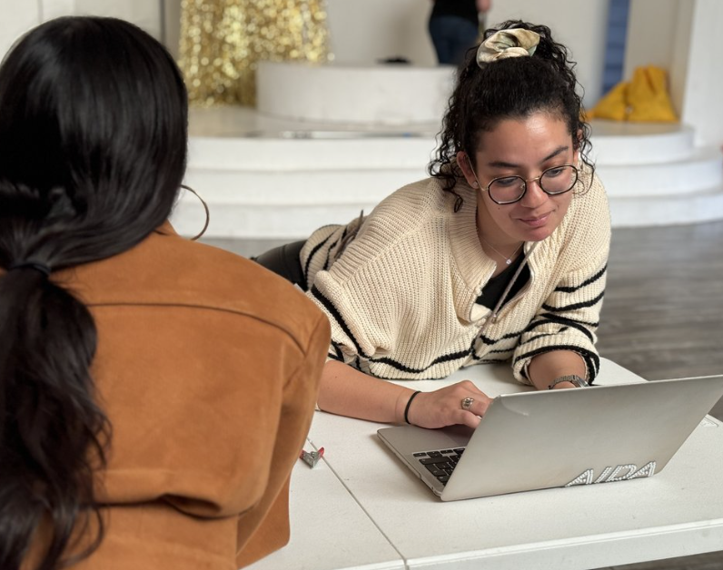 Two women sitting at a table, one with long dark hair wearing a brown jacket, the other with curly hair and glasses, working on a laptop in a bright room.