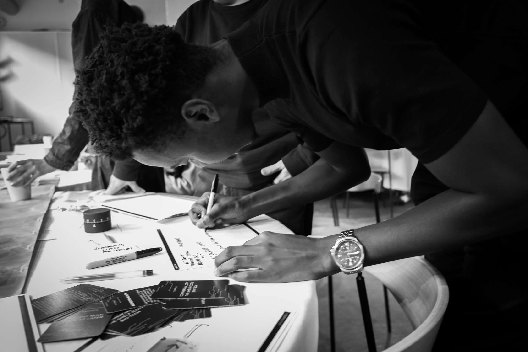 Nathanael Hondi: People gathered around a table, writing on papers with pens, in a meeting or workshop setting.