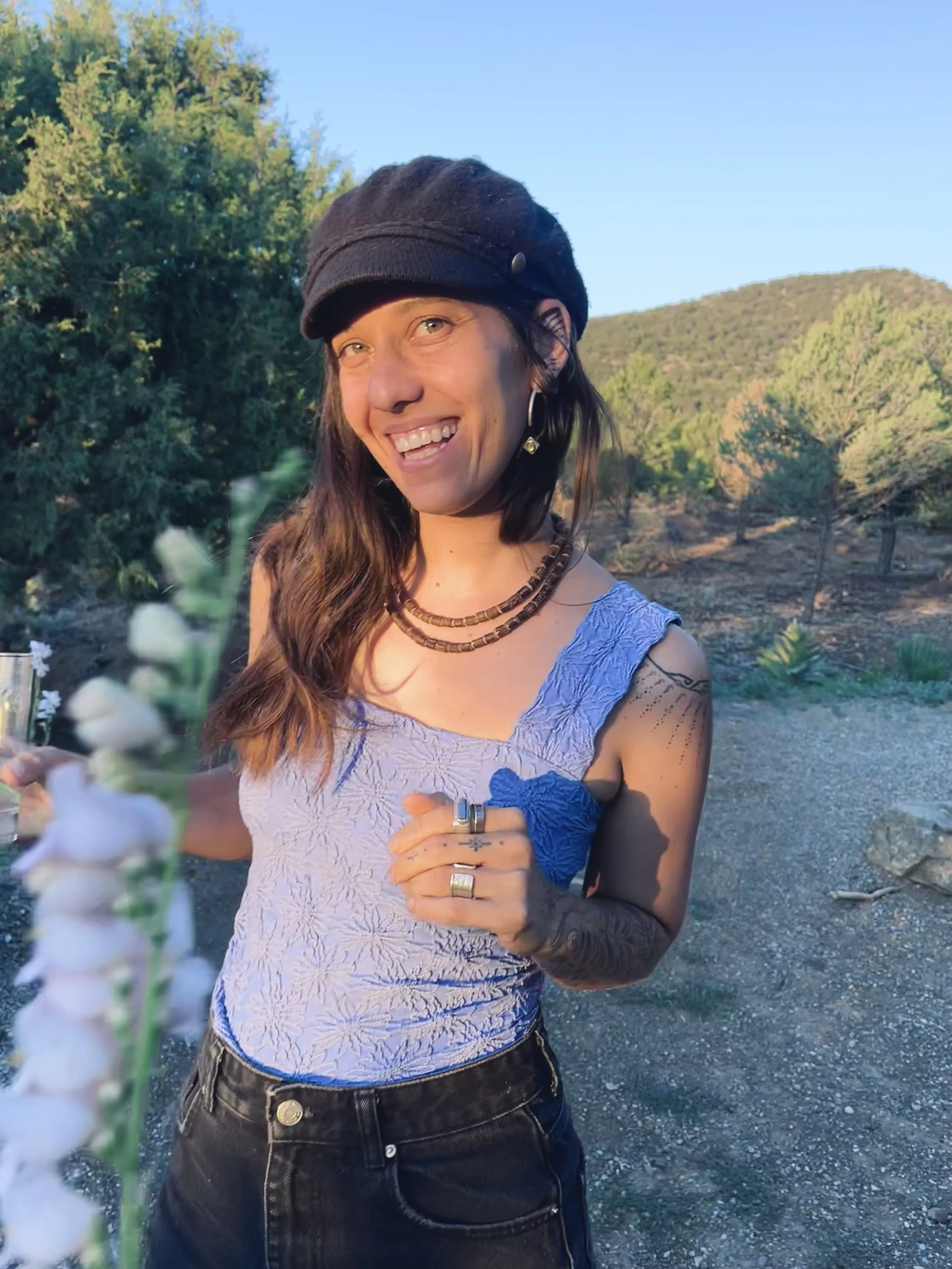 A woman smiling outdoors in sunlight, wearing a black hat, purple textured tank top, and black jeans, with tattoos on her arms, standing in front of green trees and a hillside.