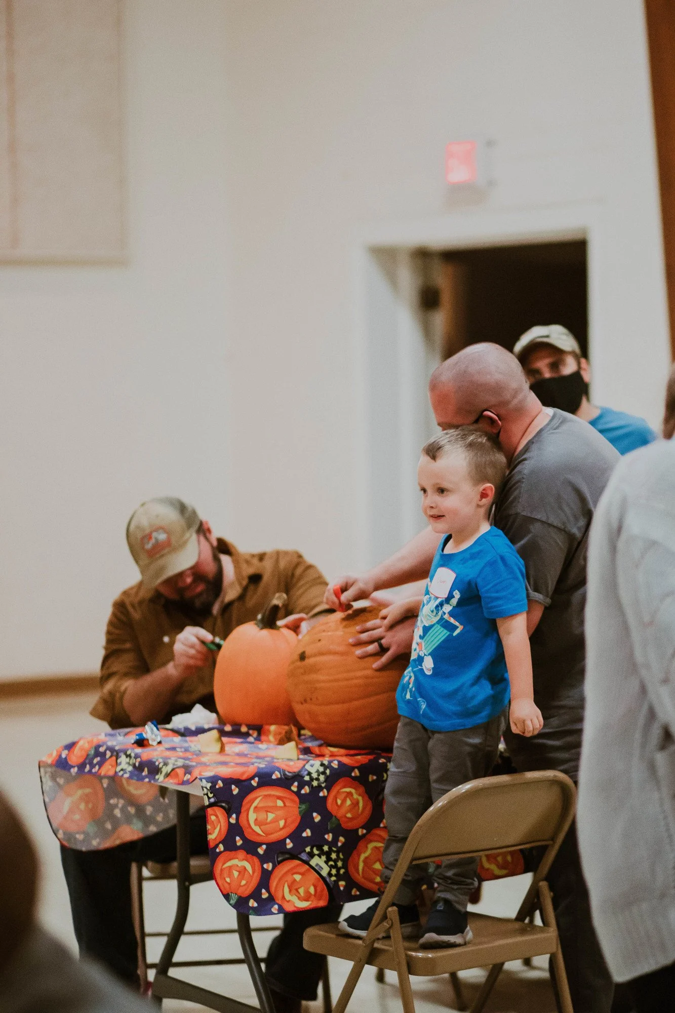 People carving pumpkins at a Halloween event, with a child standing on a chair.
