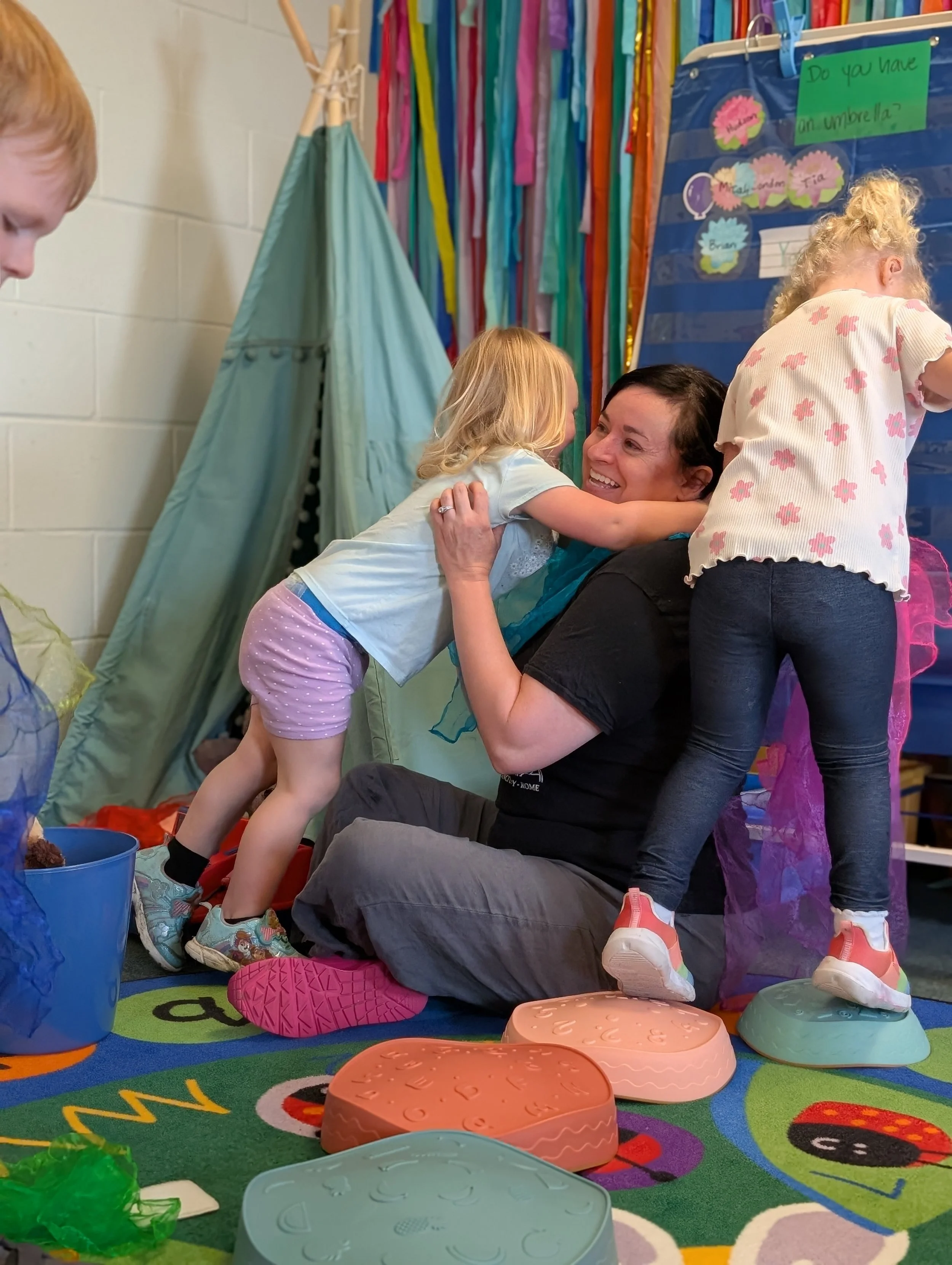 A woman sitting on the floor in a colorful room, smiling as two young girls hug her. One girl is hugging her face, and another one is standing on an animal-shaped stepping stool. The room has a bright curtain with strips of fabric, a teepee tent, and