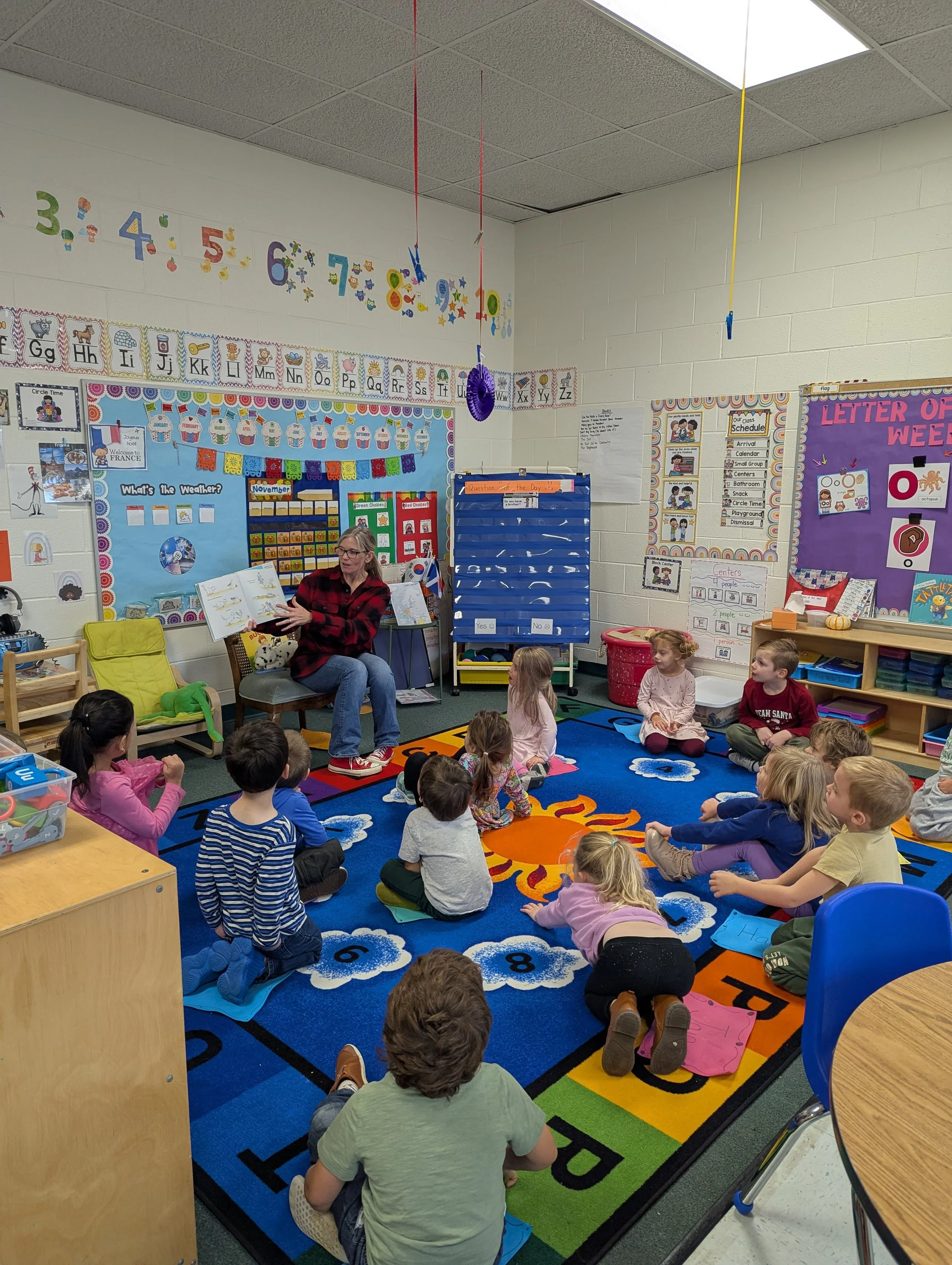 A woman reading a book to a group of young children seated on a colorful rug in a classroom with educational decorations.