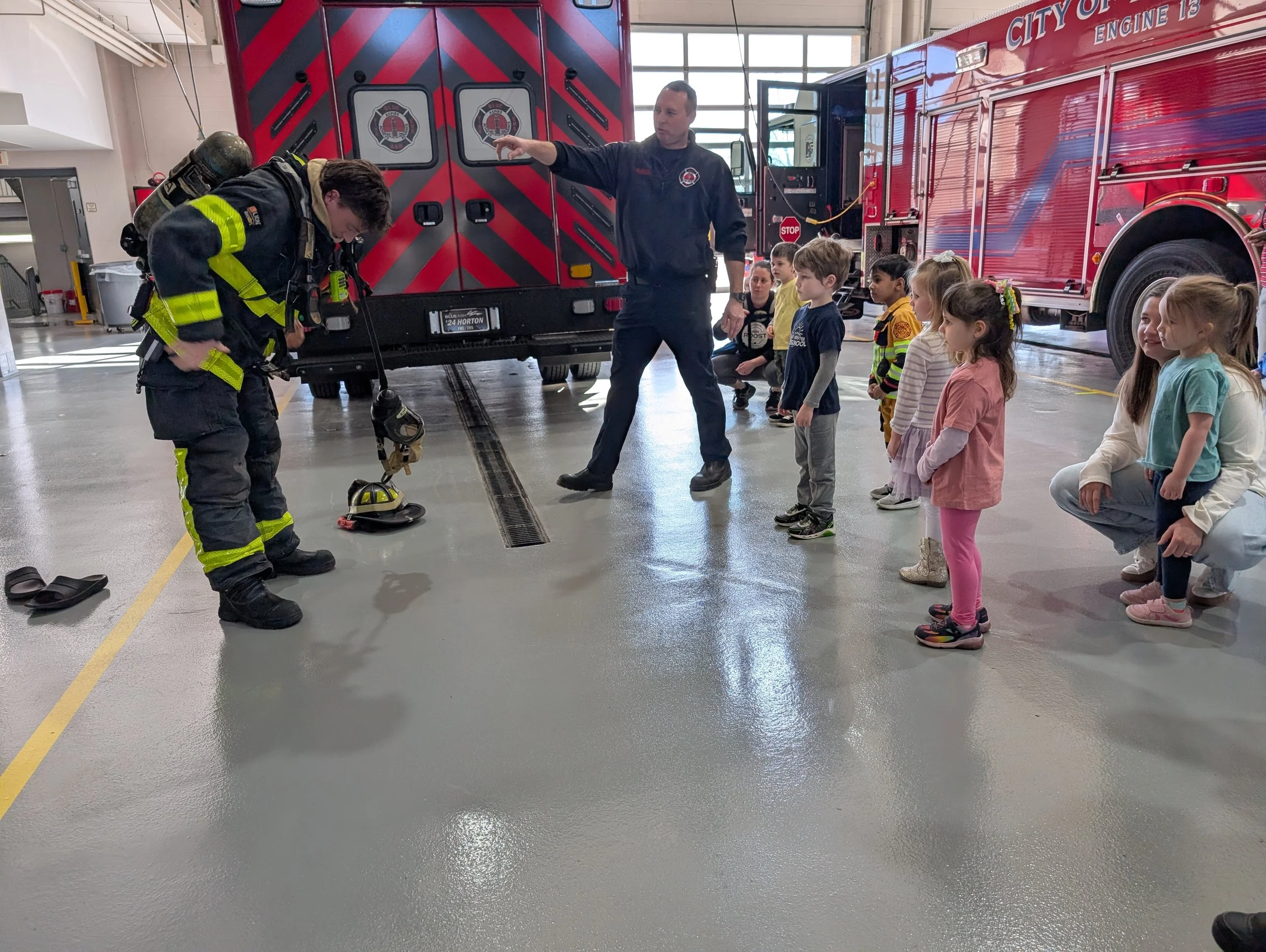 A firefighter demonstrating firefighting gear to a group of children in a fire station. The children are watching attentively as the firefighter points to the gear.