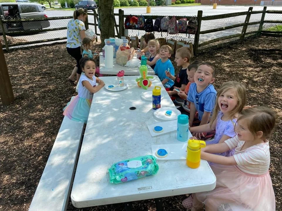 Children at a birthday party sitting at a long table outdoors, with cupcakes, water bottles, and colorful party favors, in a park area with a wooden fence and backpacks hanging nearby.