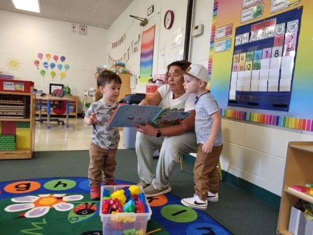 A teacher reading a book to two young children in a classroom. The children are standing next to the teacher on a colorful rug with flowers and circles. The classroom has educational posters, a whiteboard, and storage bins visible in the background.