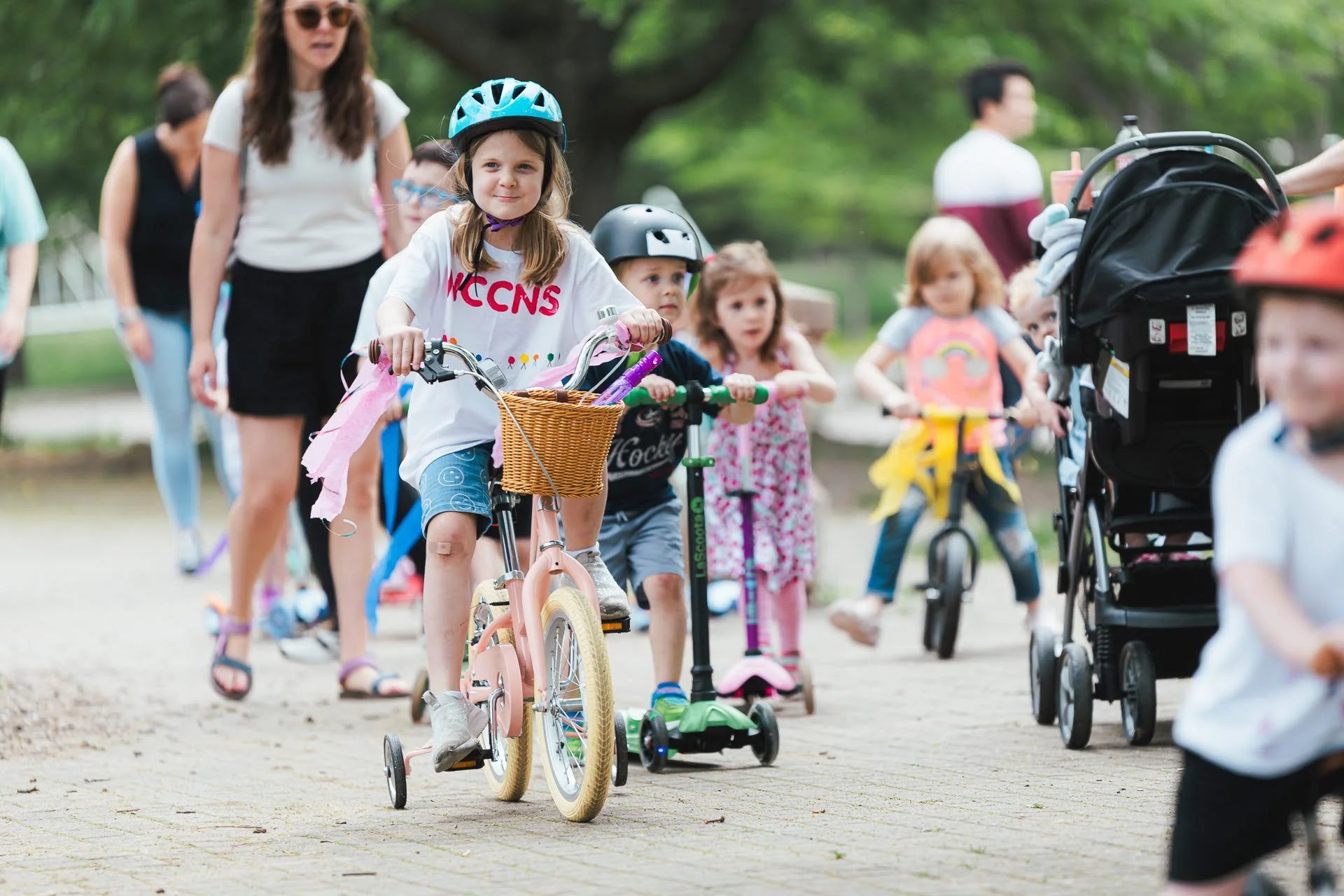 Children and a woman riding and walking with strollers, scooters, and bicycles in a park.