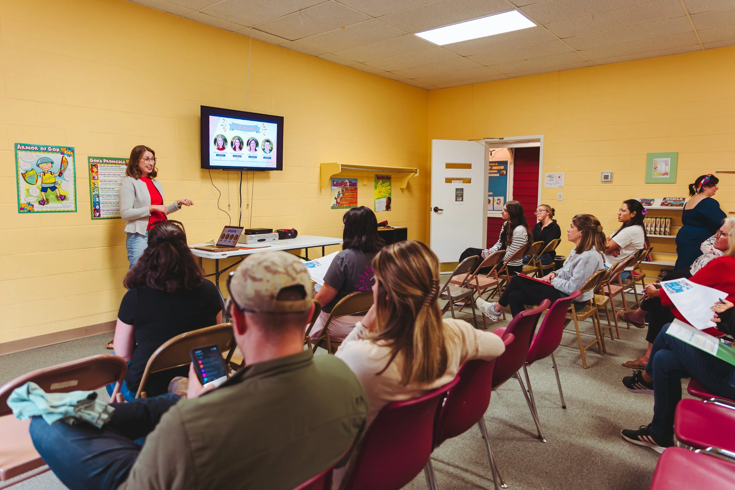 A woman gives a presentation to a group of people seated in chairs inside a classroom or community room with yellow walls. The presenter stands next to a screen displaying a slide, while the audience listens attentively, some taking notes or using th