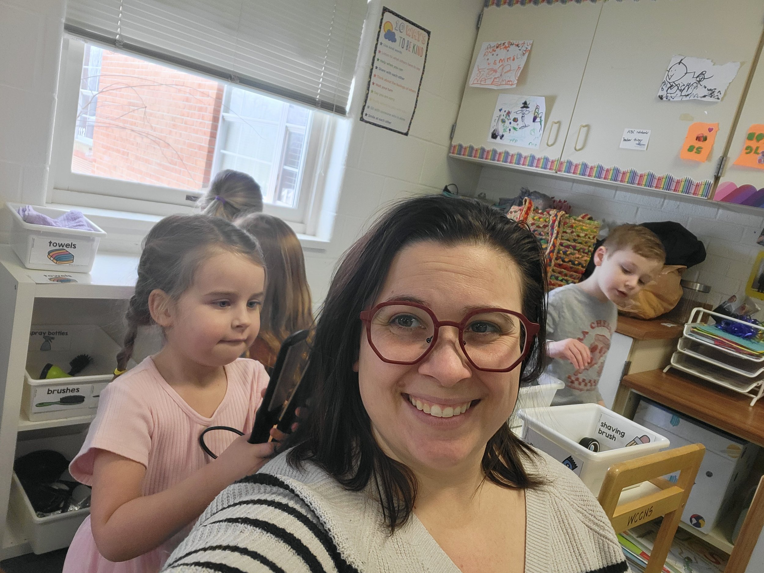 A woman with glasses taking a selfie in a preschool classroom with children in the background, some engaged in activities.