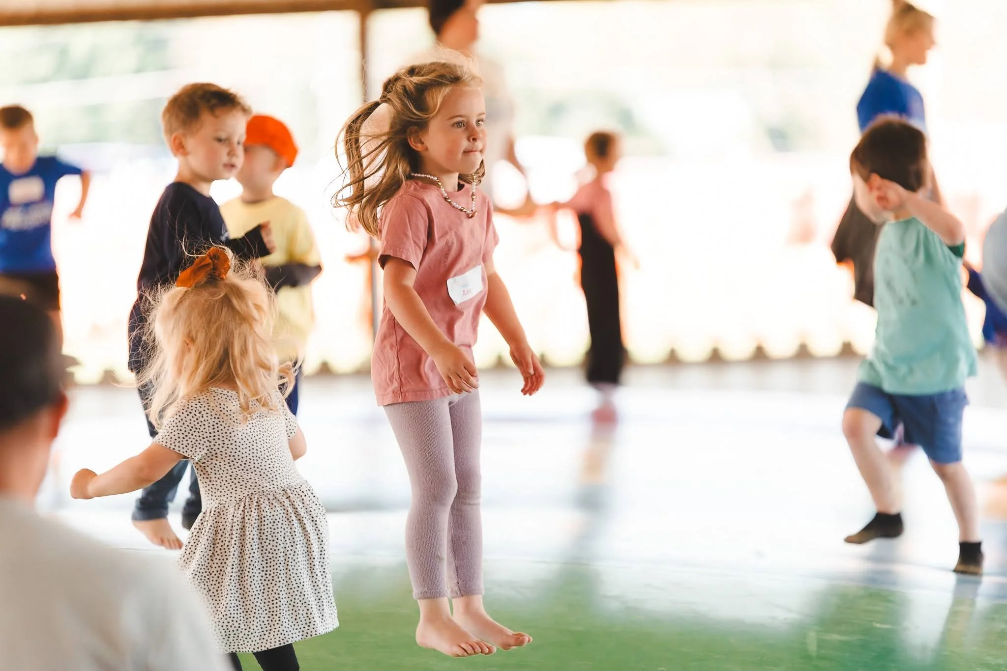 Children jumping and playing indoors, some on a green floor, during a group activity or class.