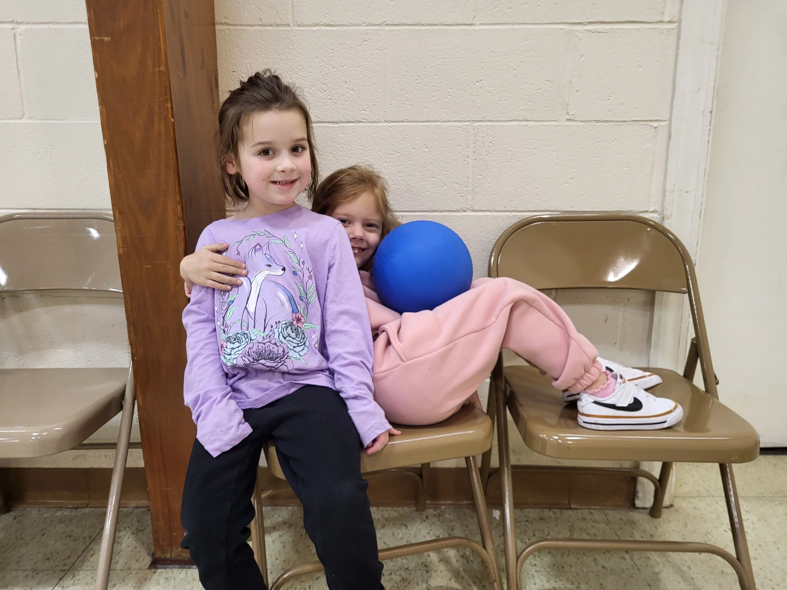 Two young girls sitting on brown chairs against a beige wall; one girl in a purple sweatshirt with a unicorn design, and the other girl in pink pajamas, holding a blue ball.