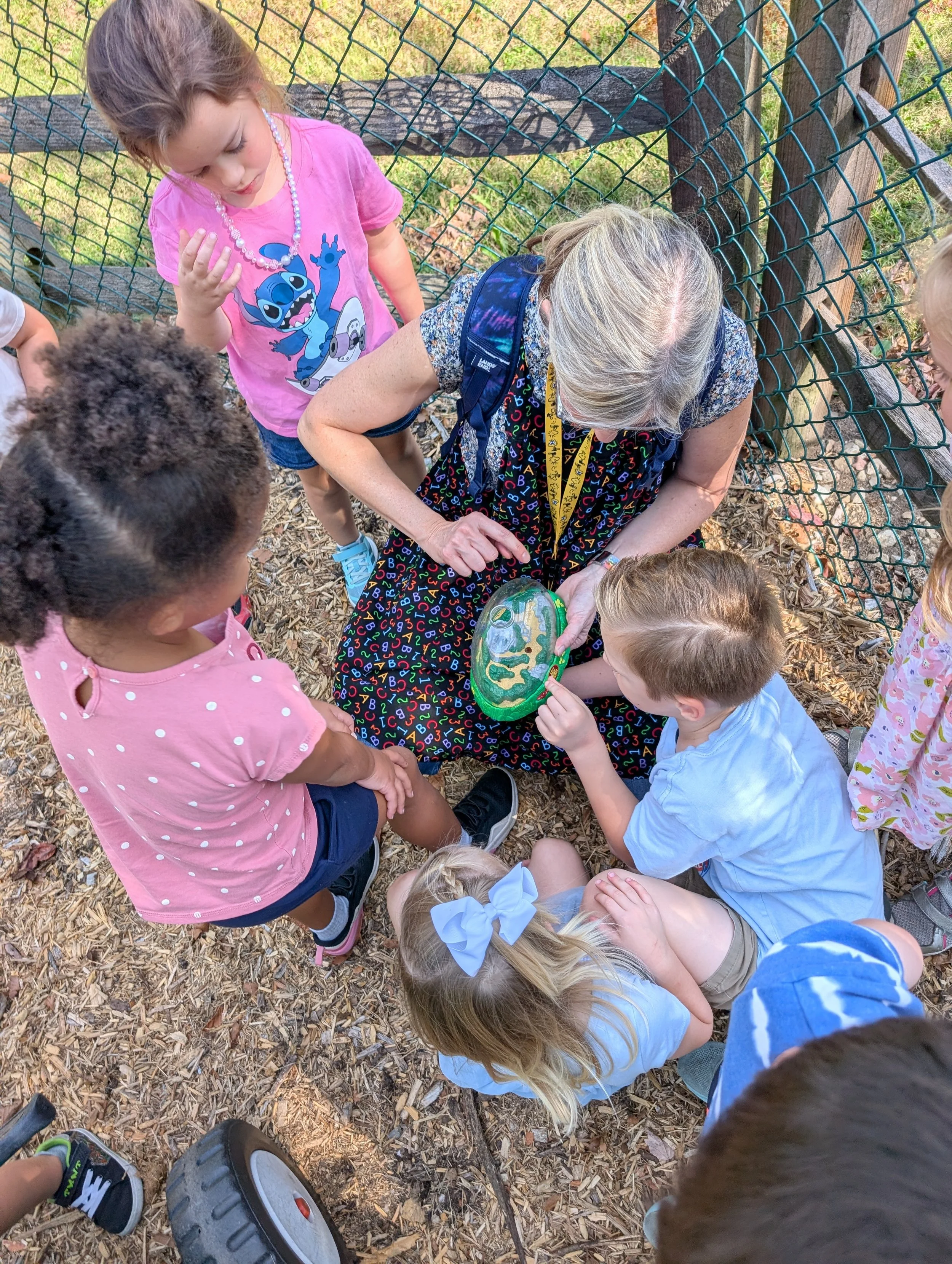 A group of children gathered around a woman sitting on the ground outdoors, showing her a painted rock with a landscape scene, with a wooden fence and grass in the background.