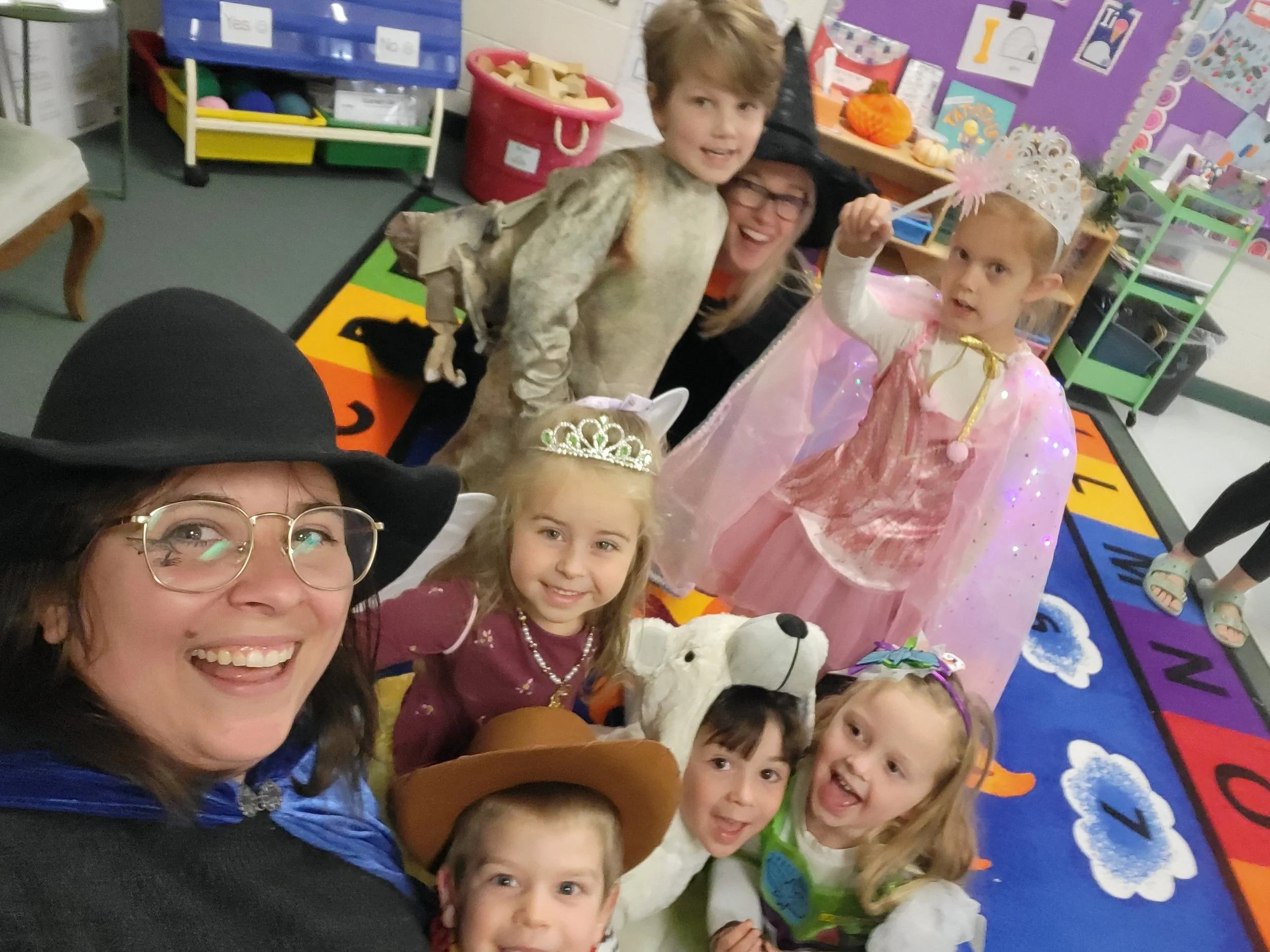 Group of children and adults dressed in costumes, some wearing princess, witch, and animal costumes, gathered in a classroom with colorful decorations and a rainbow-colored rug, celebrating a costume event.