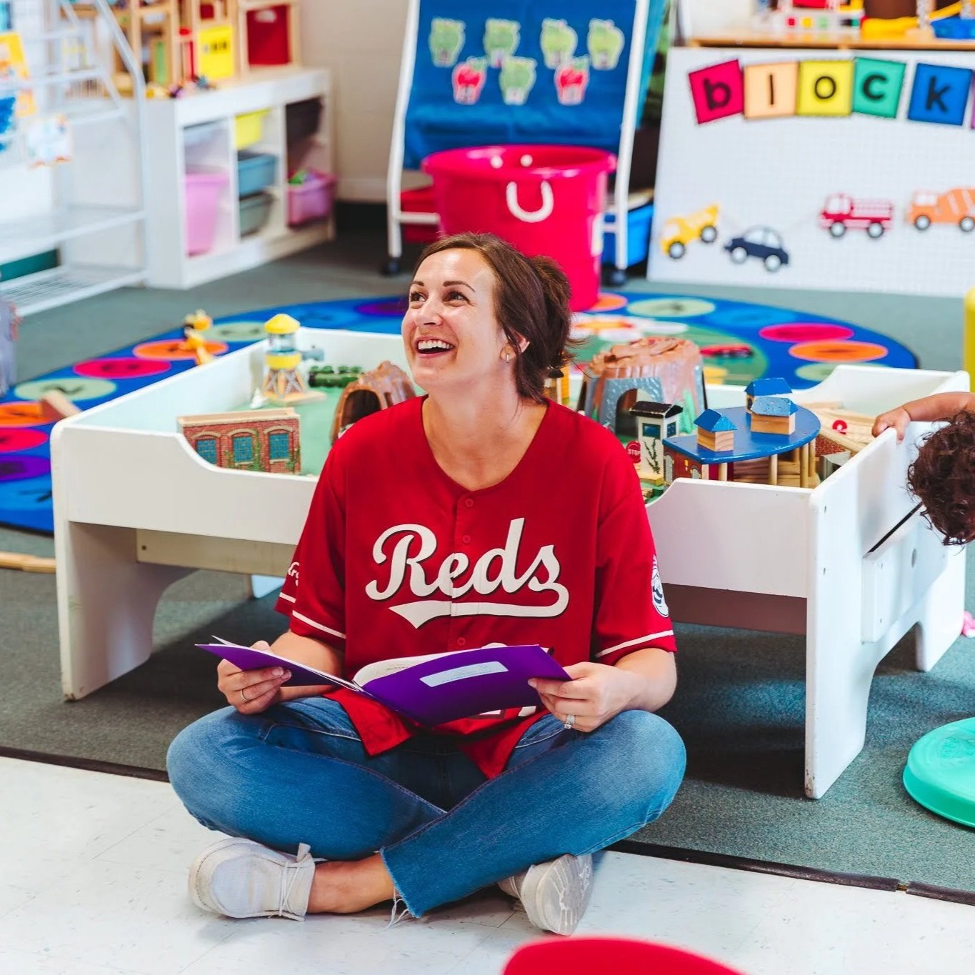 A woman sitting cross-legged on the floor in a colorful classroom, smiling and holding a purple folder, surrounded by a toy train table, colorful teaching materials, and decorations.