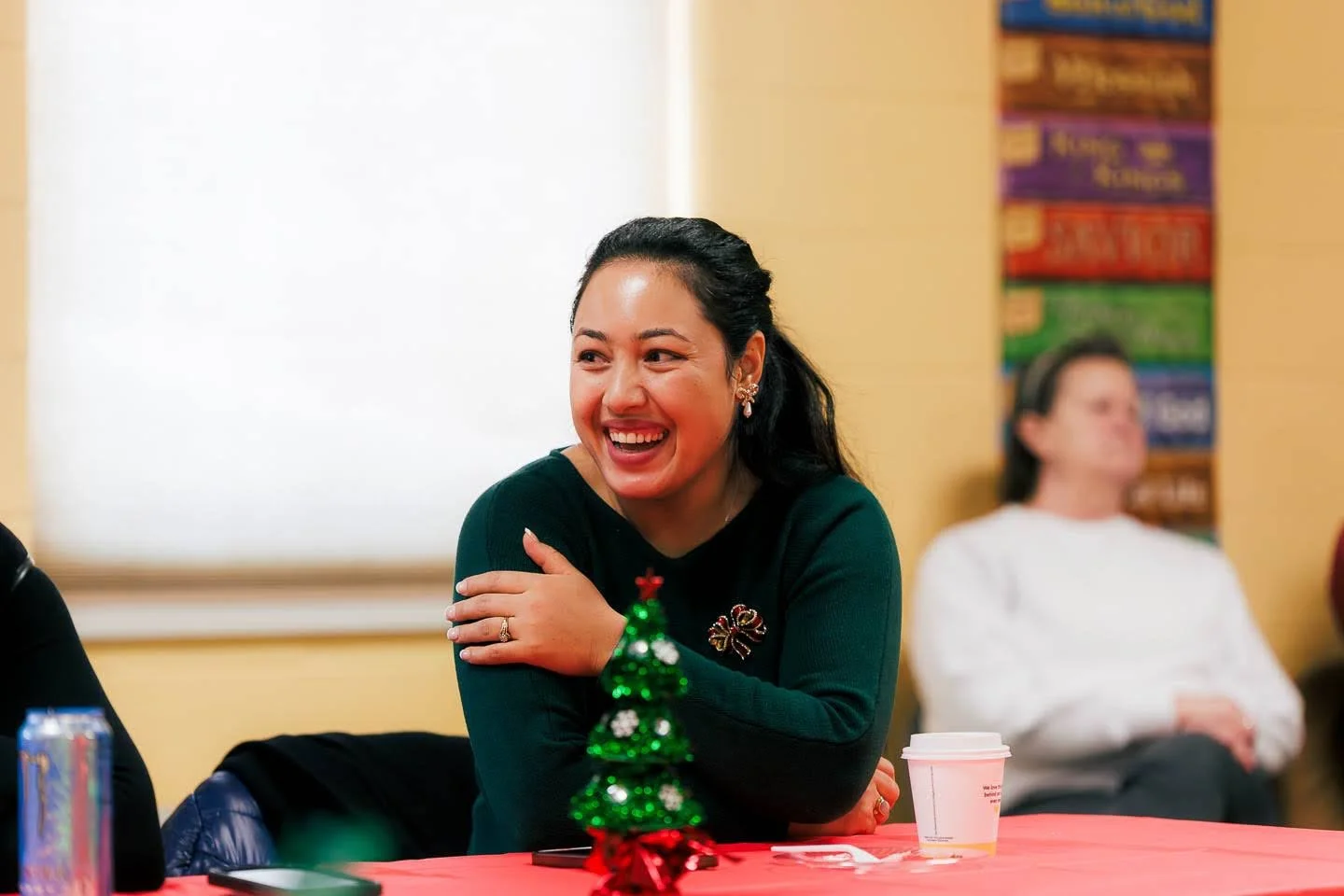 A woman with dark hair smiling and holding her shoulder, sitting at a table with a small festive green Christmas tree decoration and a white coffee cup in front of her during a holiday gathering.