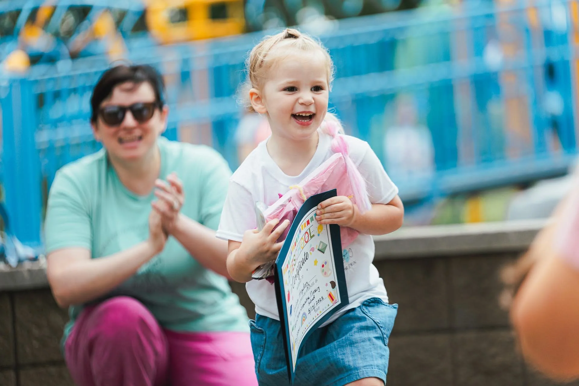 A young girl with blonde hair in a braid, smiling and holding a certificate, running in front of a woman clapping. They are outdoors near a blue fence, in a park or playground setting.