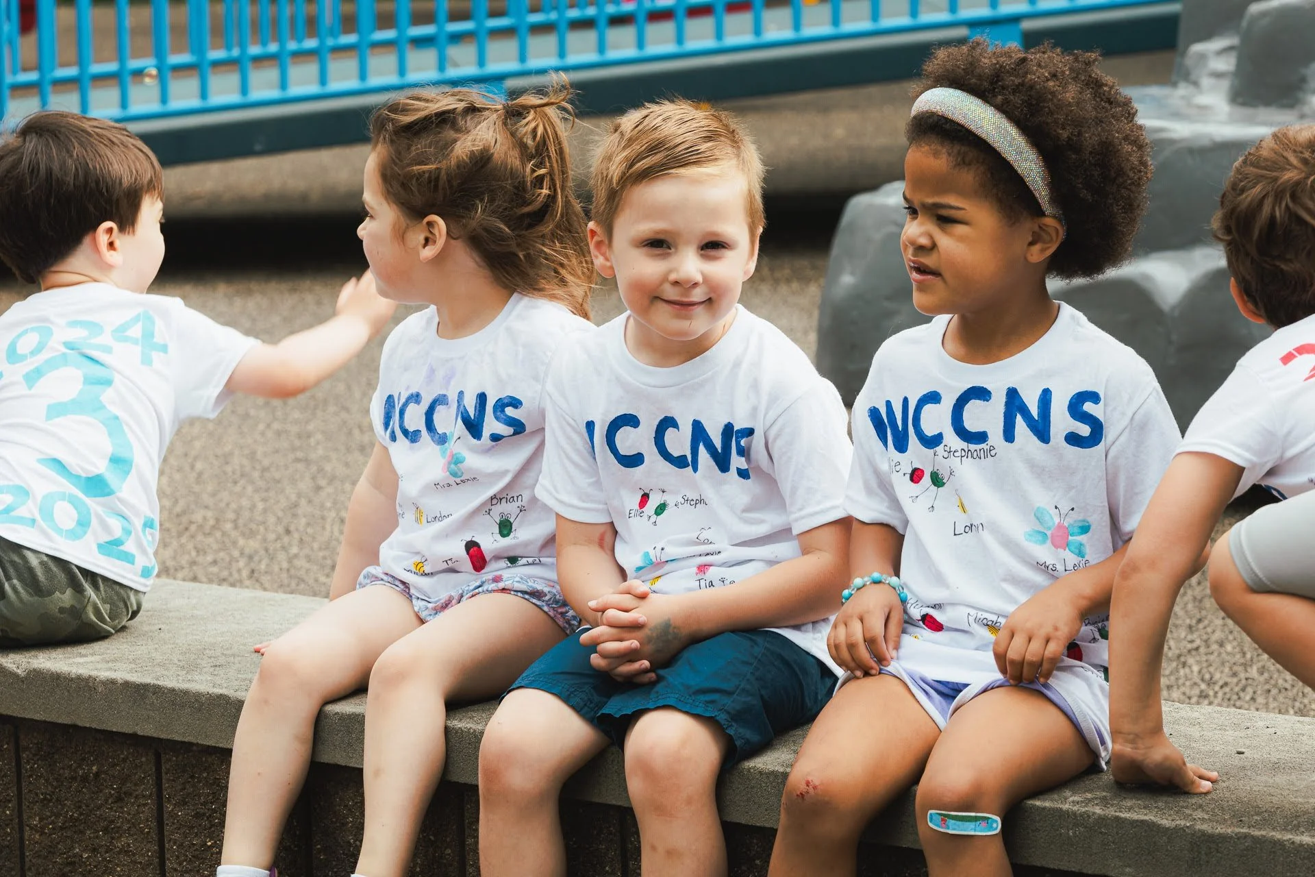 Four children sitting on a concrete ledge outdoors, wearing white T-shirts with blue writing, waiting at a public event or gathering.