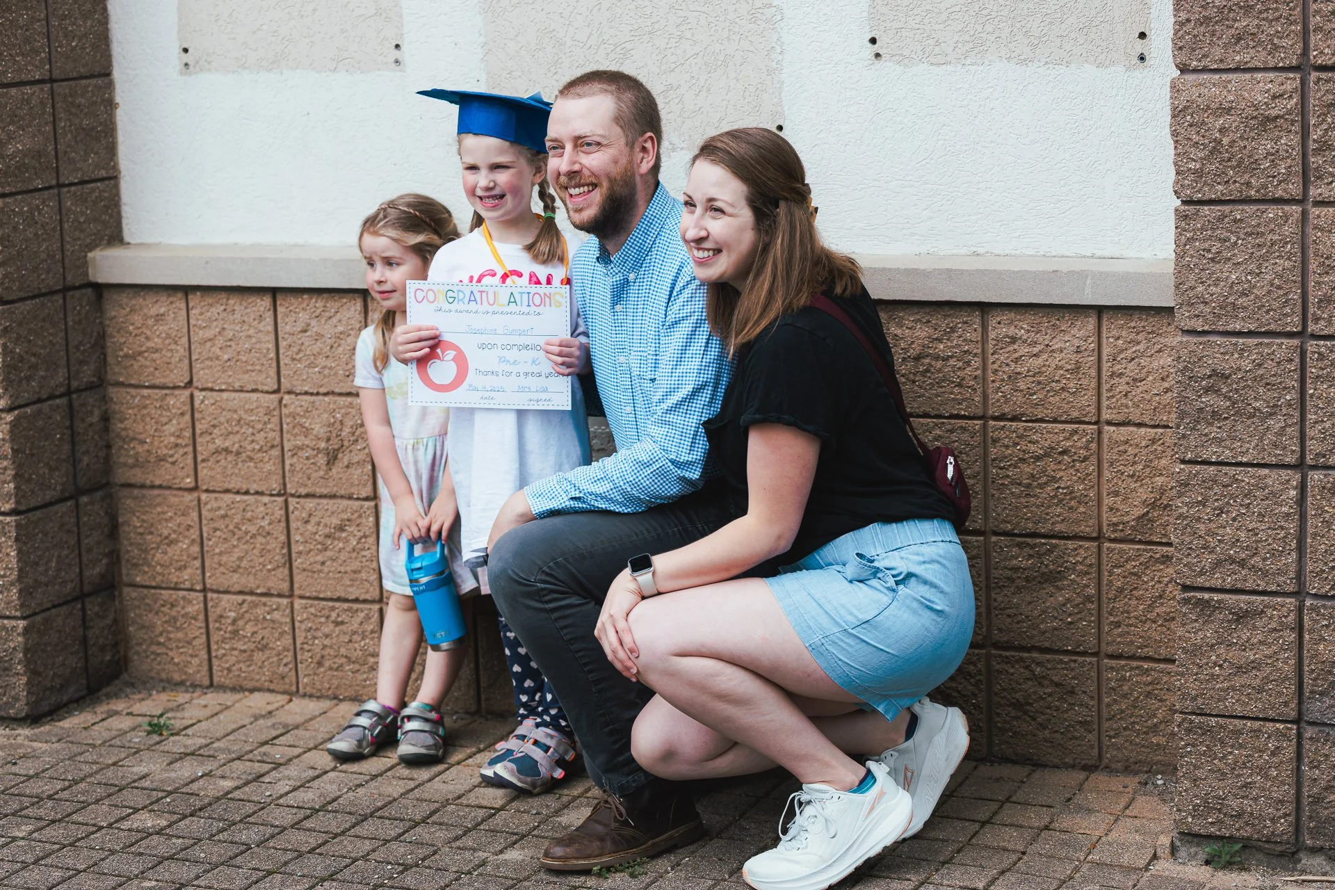 A family celebrating a child's graduation outdoors, with a young girl wearing a blue cap holding a congratulatory certificate, their parents and two young girls smiling near a brick wall.