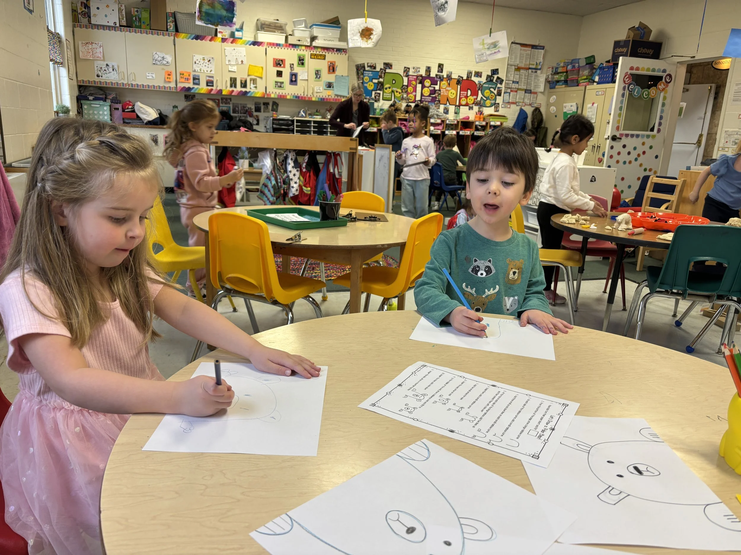 Two young children sitting at a round table drawing pictures of teddy bears in a colorful classroom with other children and teachers in the background.