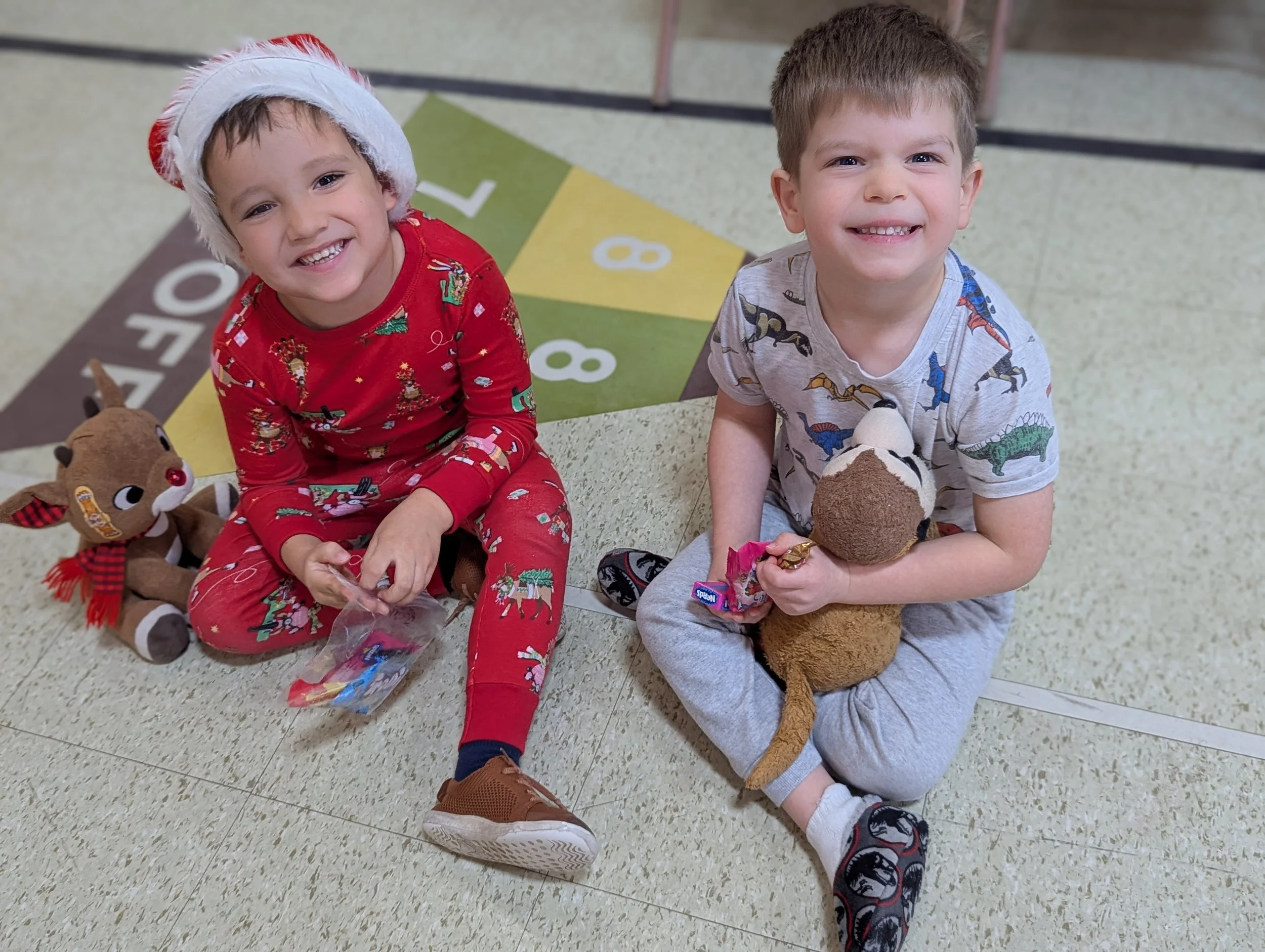 Two young boys sitting on the floor, smiling at the camera. One is wearing a red Christmas-themed outfit and a Santa hat, holding a small bag of treats. The other is in a gray T-shirt with dinosaur prints, holding a stuffed animal and a candy. They a