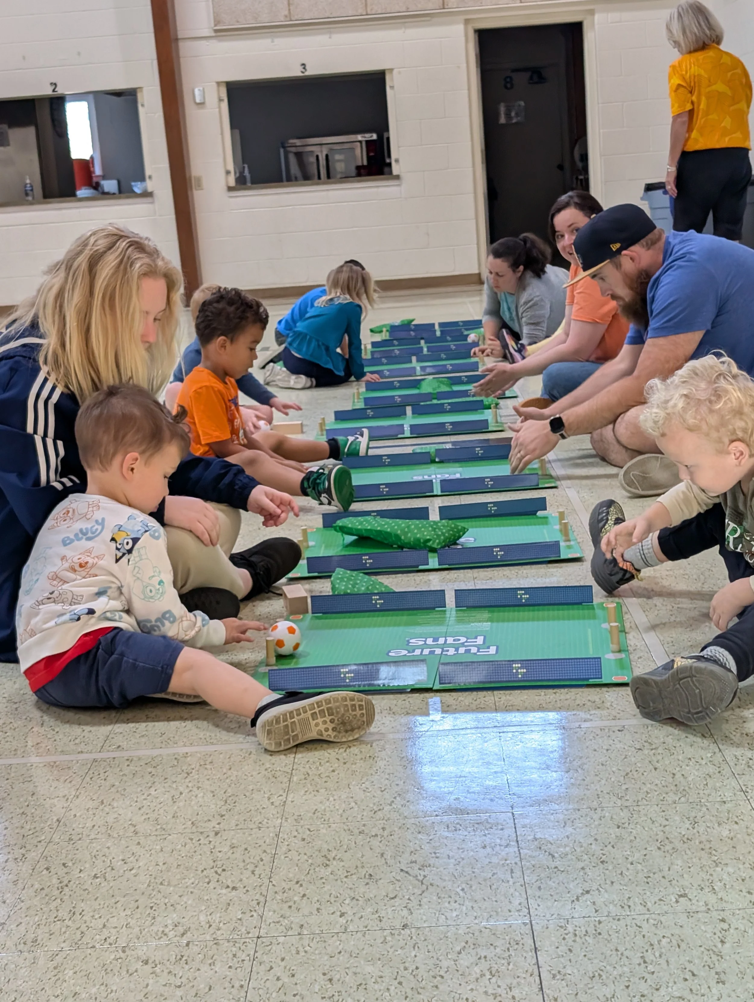 Children and adults sitting cross-legged on the floor in a line, playing a miniature hockey game with small paddles and balls, in an indoor community center or gym.