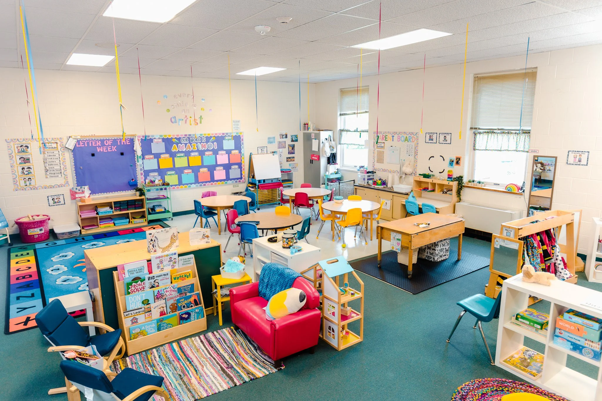 Colorful preschool classroom with small tables and chairs, educational posters, books, and various learning activities.