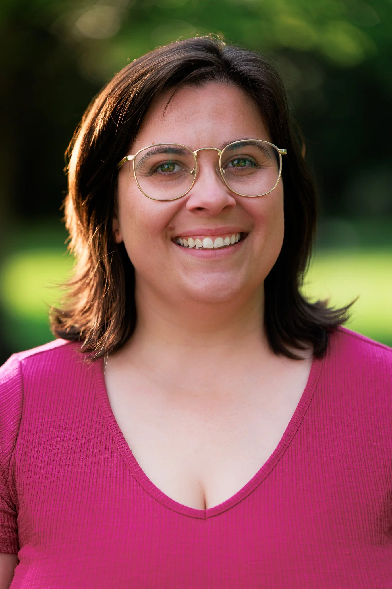 Lexie Lenavitt with shoulder-length brown hair, wearing glasses and a pink top, smiling outdoors with a blurred green background.