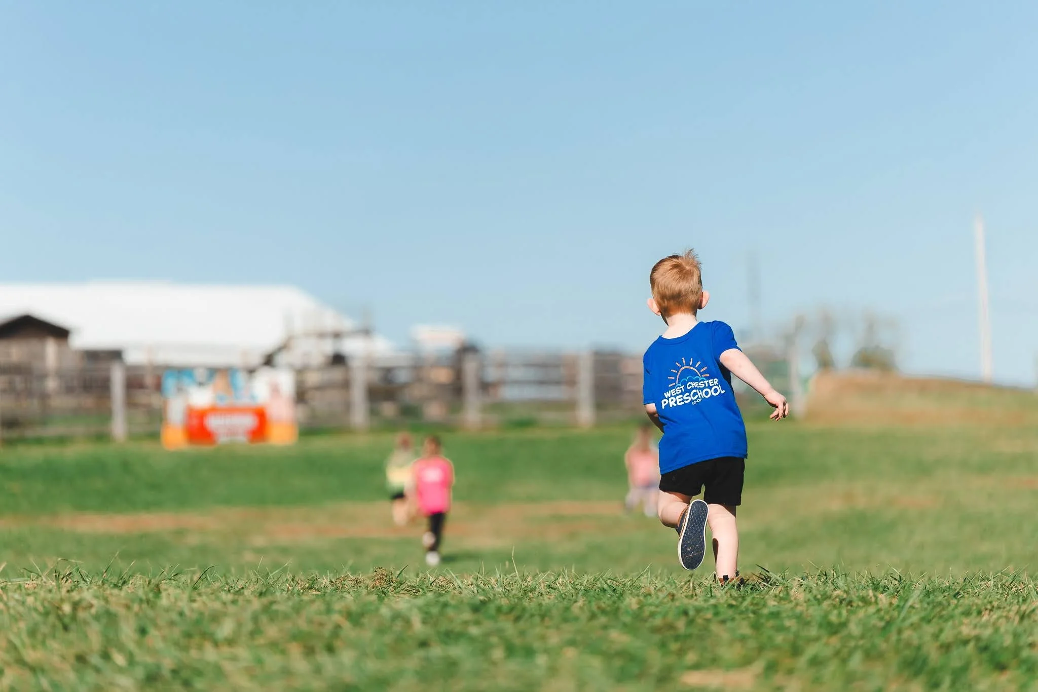 Child running on grassy field during outdoor event with others in the background, wearing a blue t-shirt that says 'West Chester Preschool' on the back.