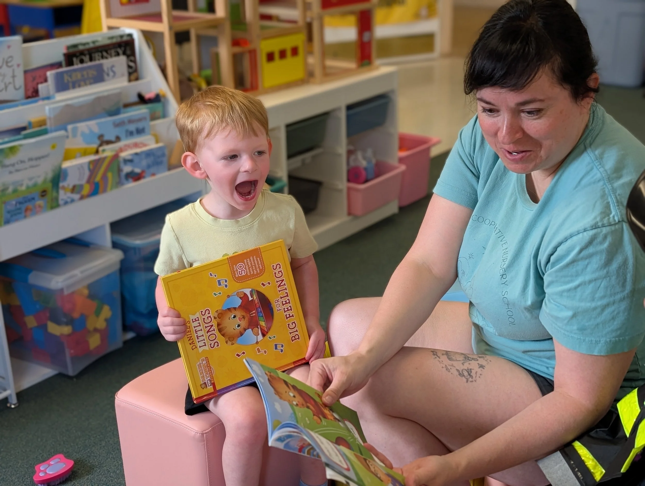Young child sitting on a pink ottoman, excitedly reading a children's book titled 'Little Singers for Big Feelings' while a woman, possibly a teacher or caregiver, sits beside them holding another children's book, in a colorful library or playroom se