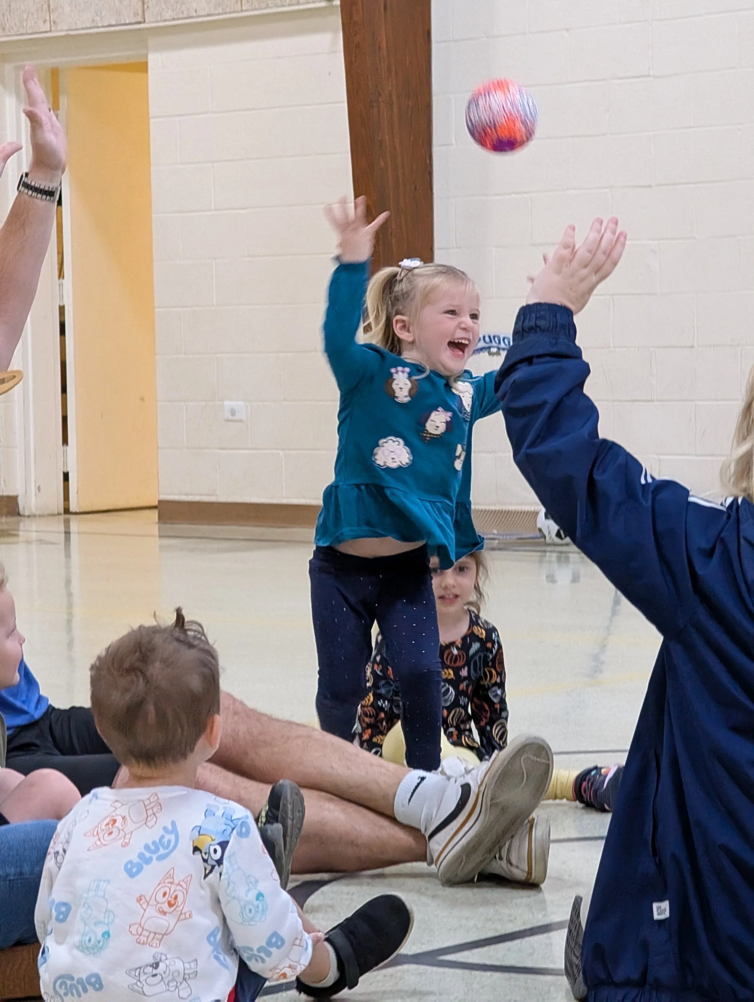 A group of children and adults sitting on the floor in a gymnasium, with a young girl in a blue shirt jumping and reaching for a colorful ball thrown into the air.