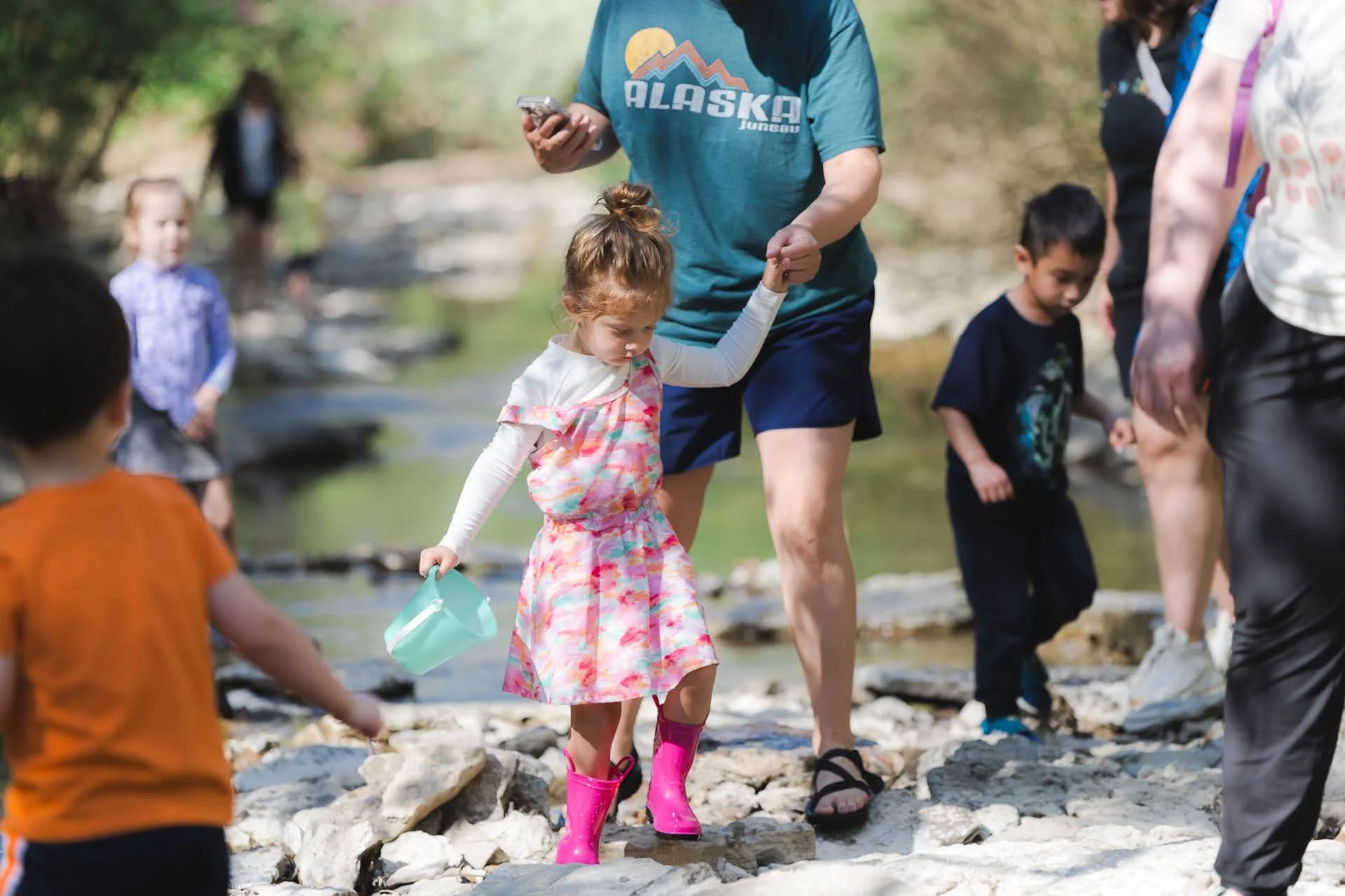 Children and adults hiking across rocky stream in forest, some children wearing boots, one girl in pink boots with a small blue bucket, adults guiding children.