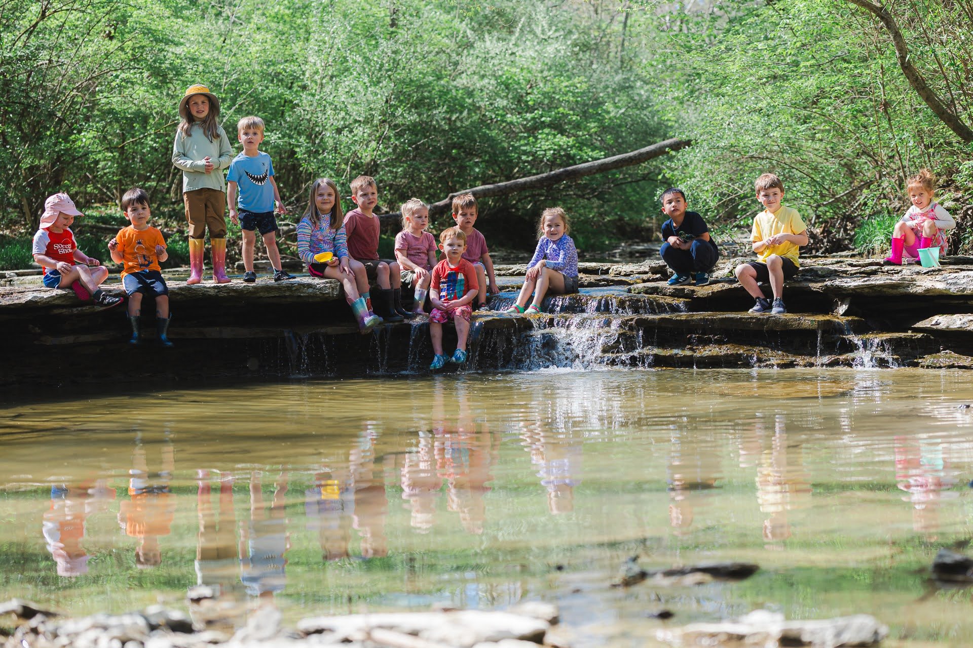 Group of children sitting and playing near a small waterfall and stream in a lush green forest.