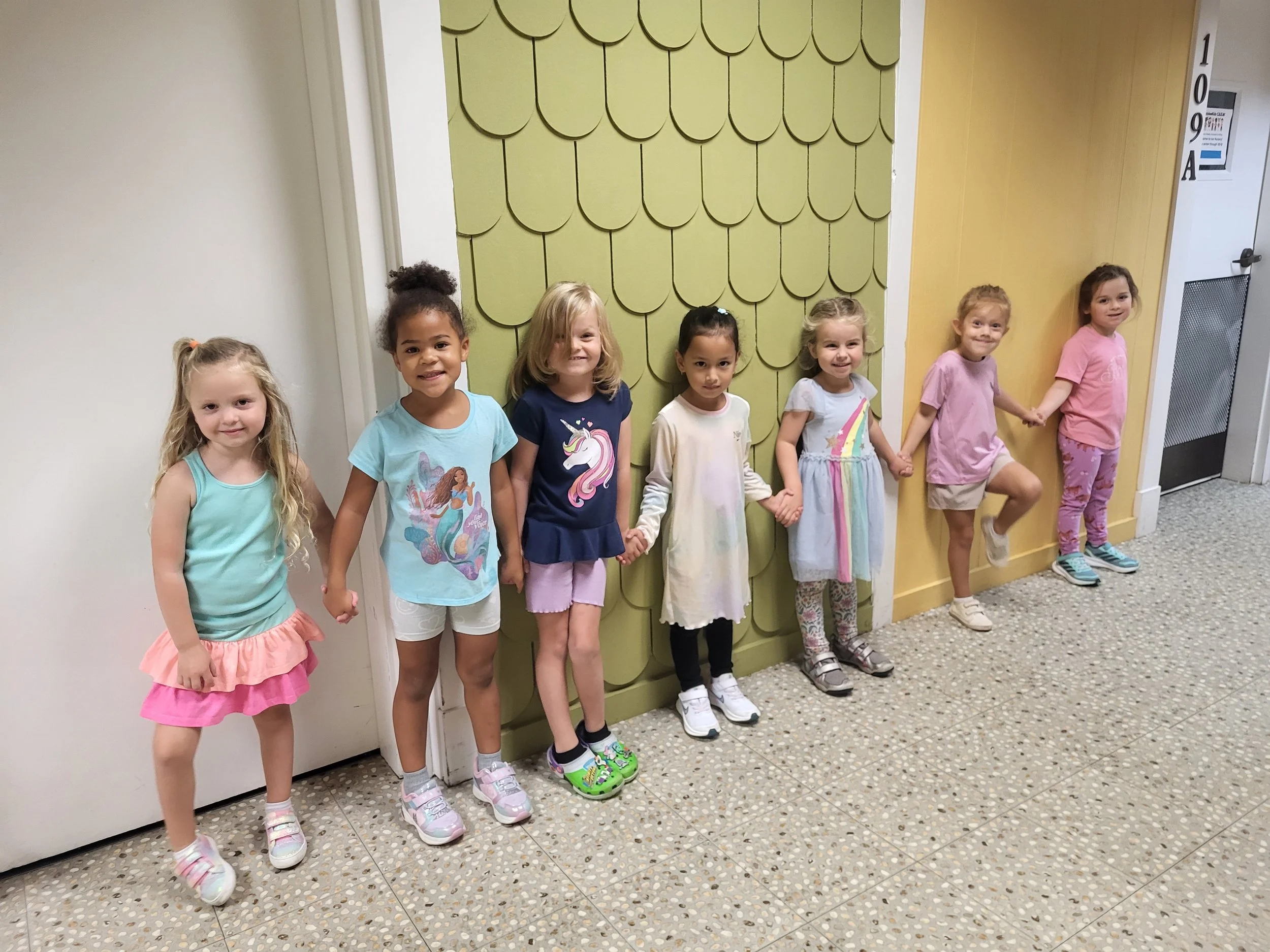 Seven young girls holding hands and standing in a line against a decorative wall. They are smiling and dressed in colorful casual clothing.