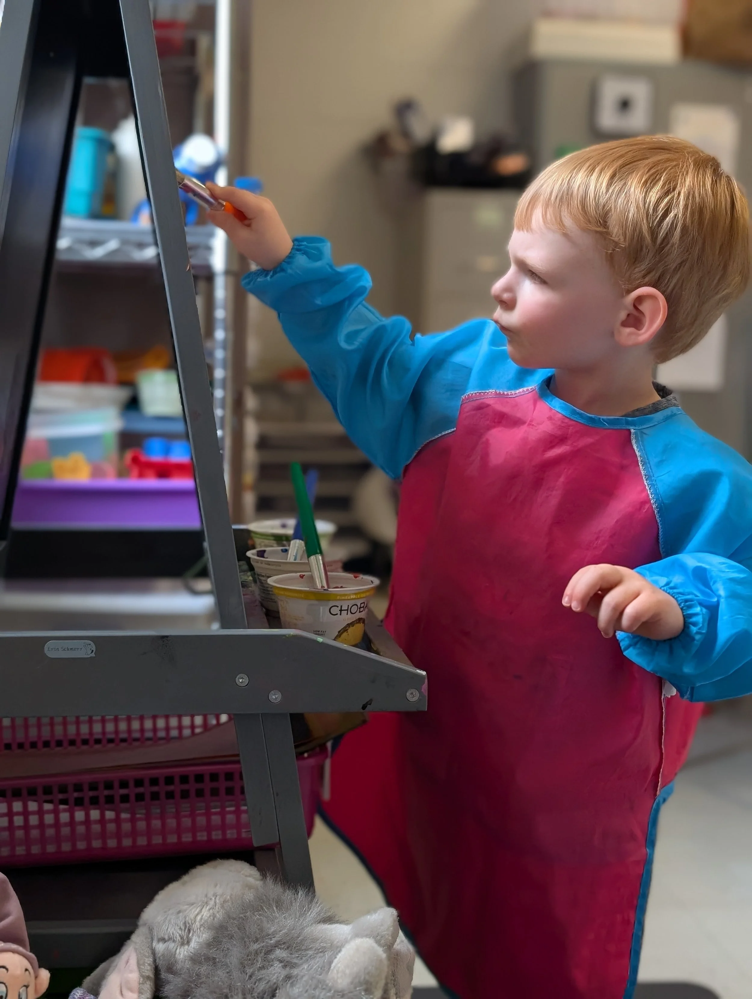 A young child with red hair wearing a pink and blue apron is reaching out with a spoon or stick in front of a standing mirror, while inside a room with storage shelves and containers.