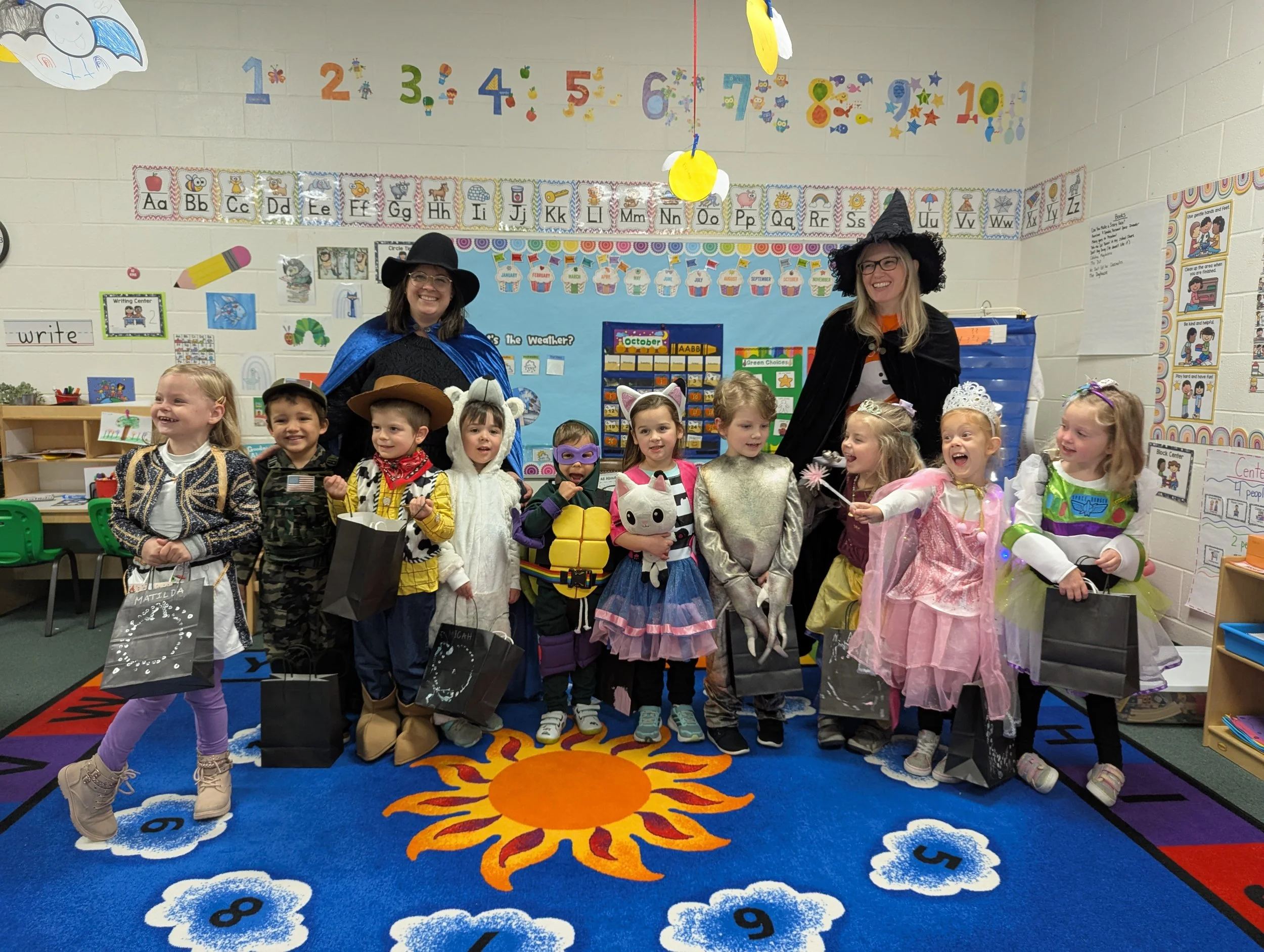 Children in costumes standing in a classroom with two women dressed as witches, celebrating Halloween and holding gift bags.