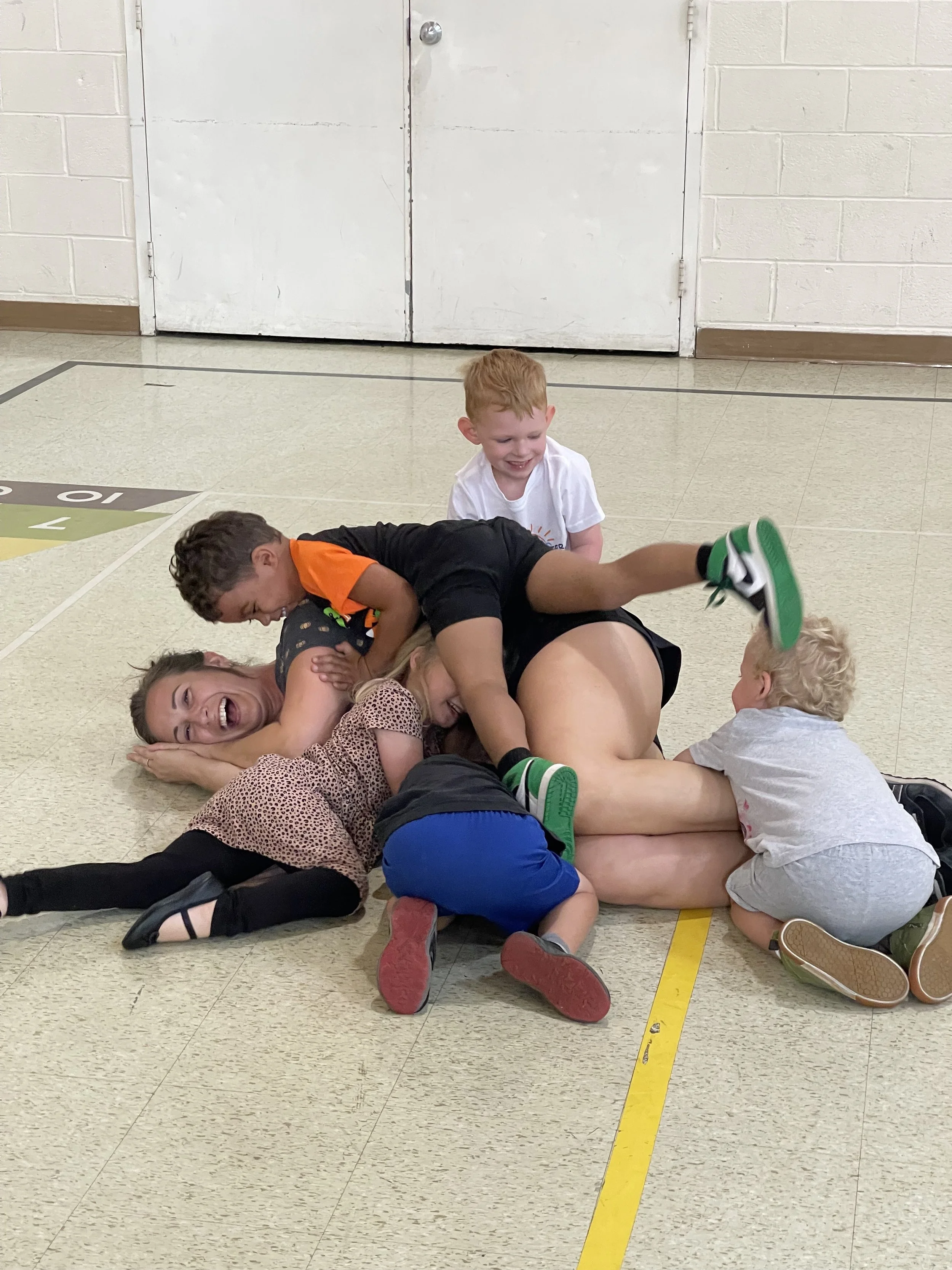 Children and an adult playfully pile on each other on a gymnasium floor, laughing and smiling.