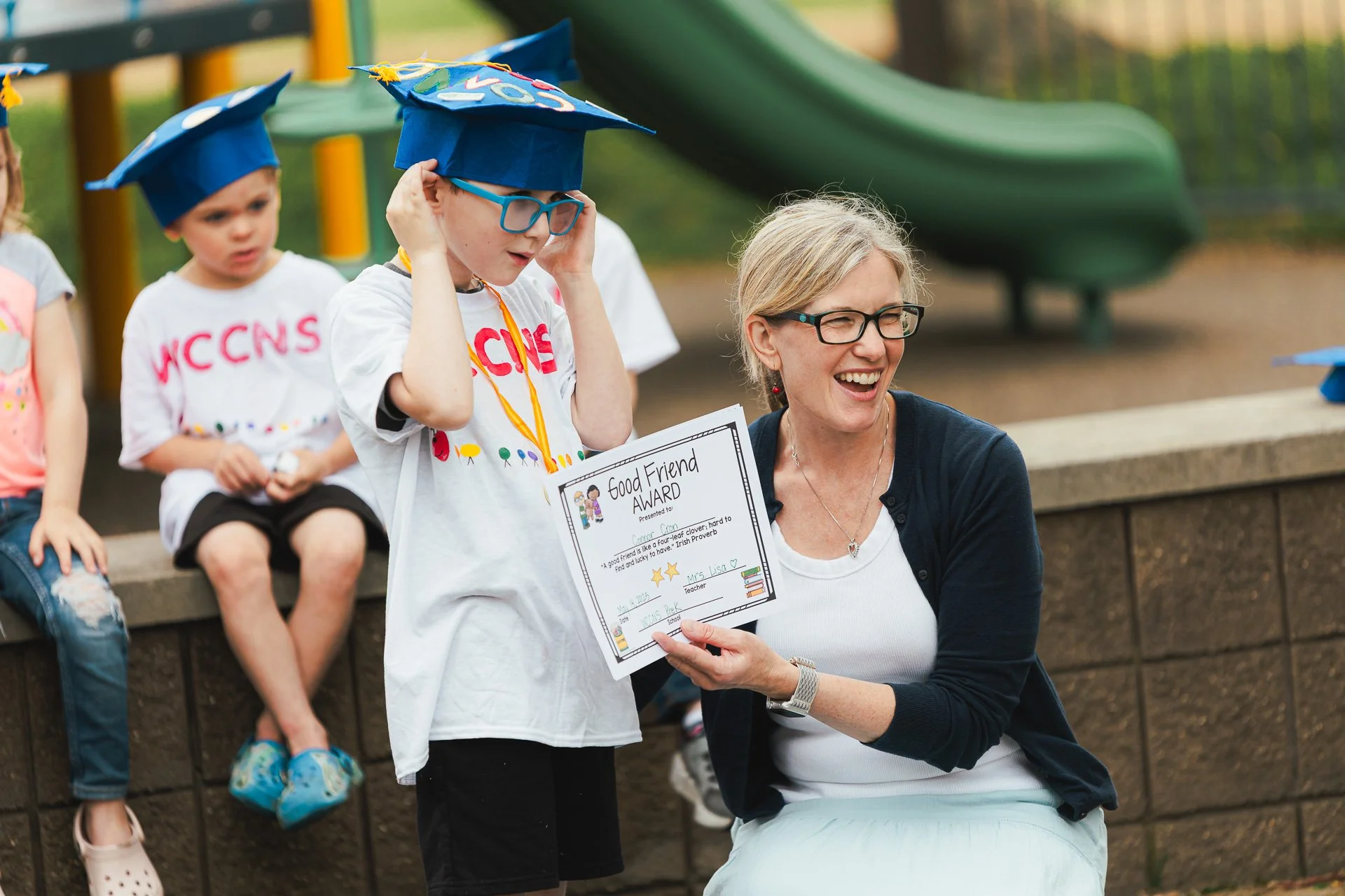 A smiling teacher awards a young student with a certificate for being a good friend at an outdoor school event. The boy is wearing a graduation cap and glasses, while other children sit nearby.