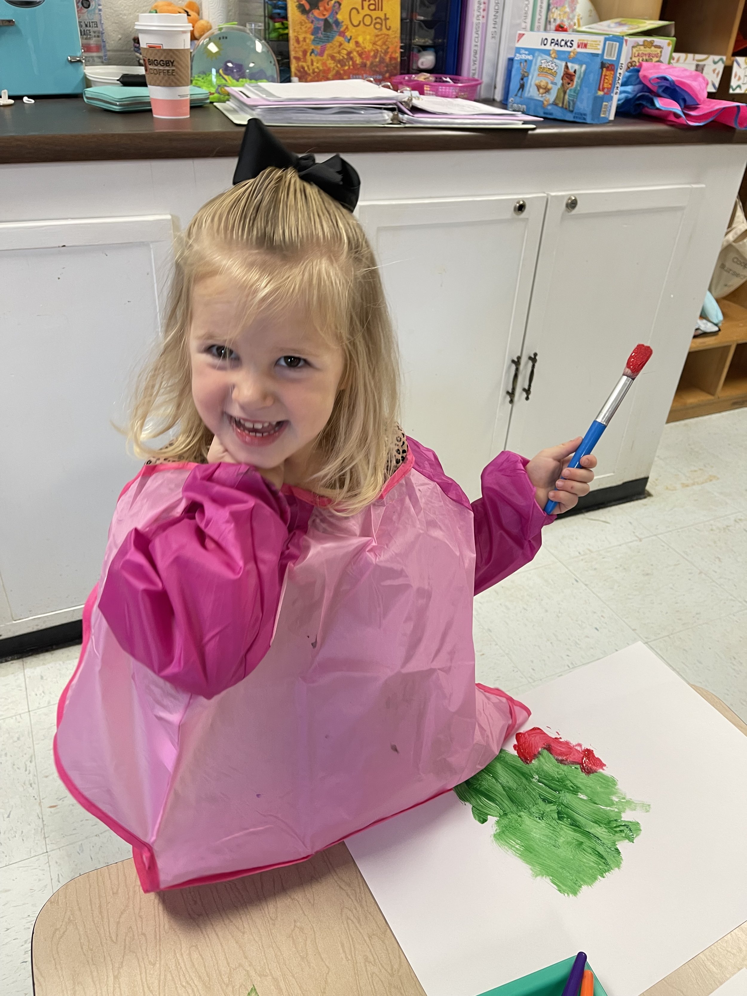 A young girl with blonde hair, wearing a pink smock with puffy sleeves and a black bow in her hair, is smiling and holding a paintbrush with red paint on it. She is painting a green Christmas tree with red decorations on a large sheet of white paper 