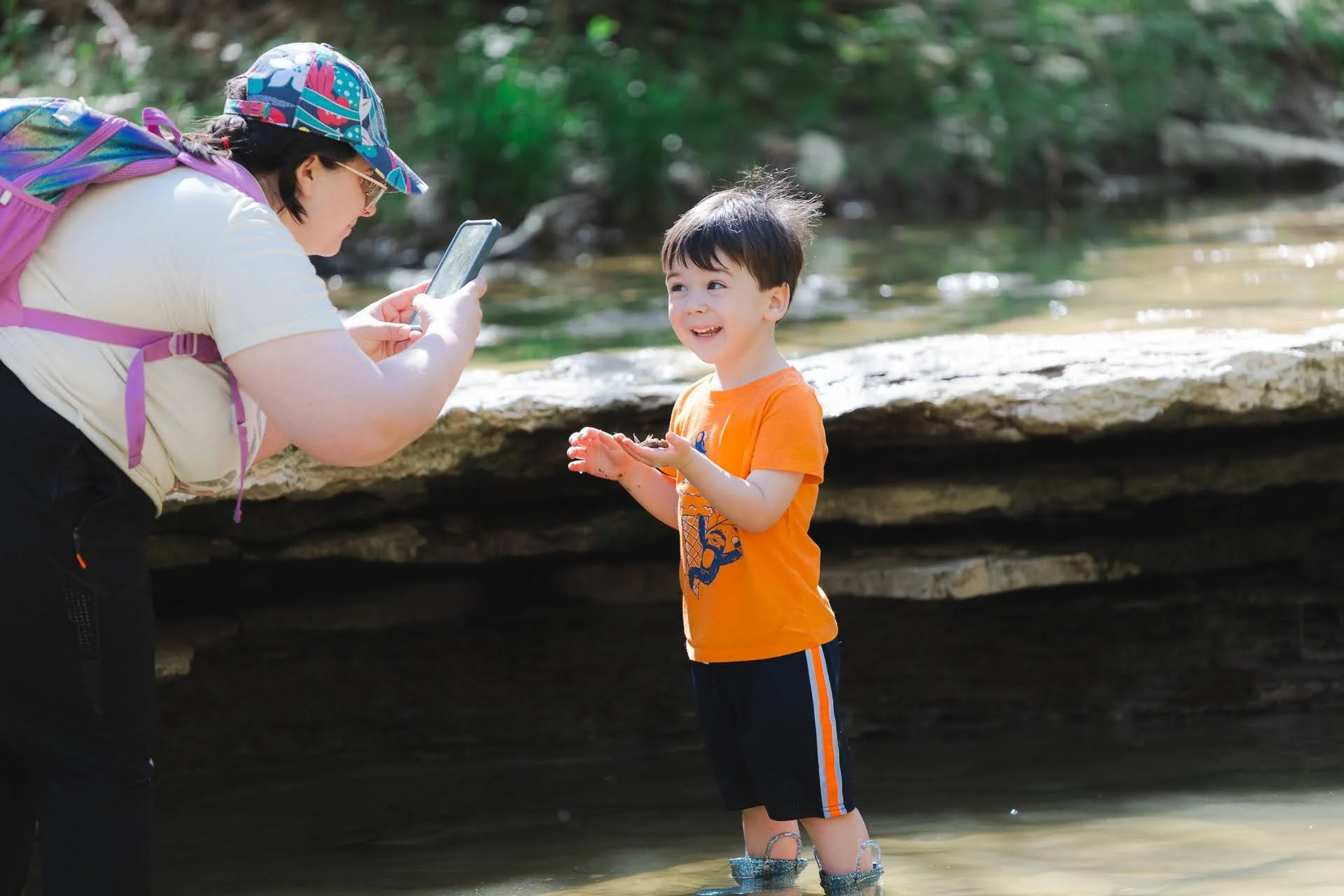 A young boy standing in a shallow creek with muddy feet, smiling at a woman who is taking his photo. The woman is wearing a colorful hat, sunglasses, and a backpack.