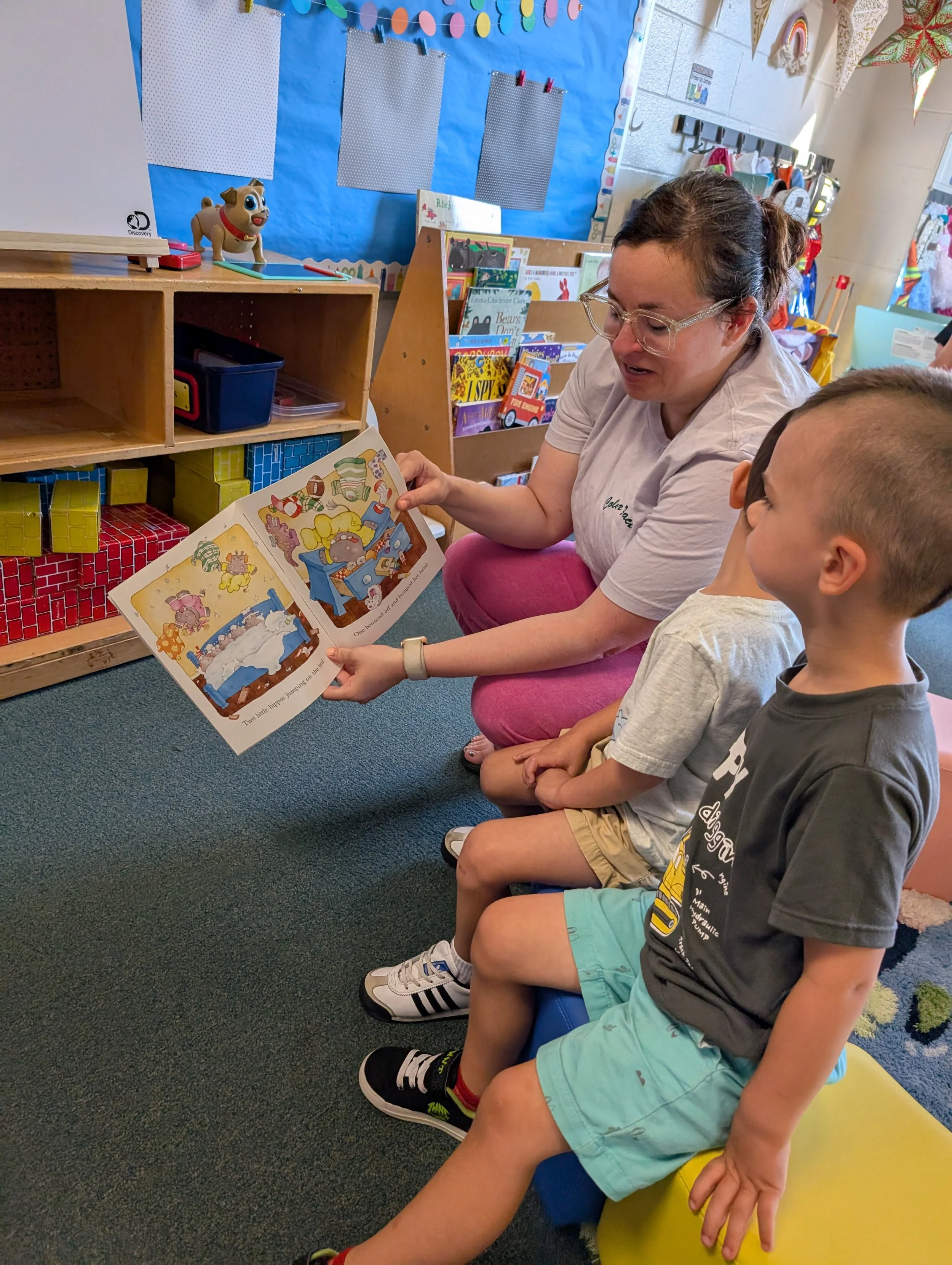 A woman reading a children's book with two young boys in a classroom setting.