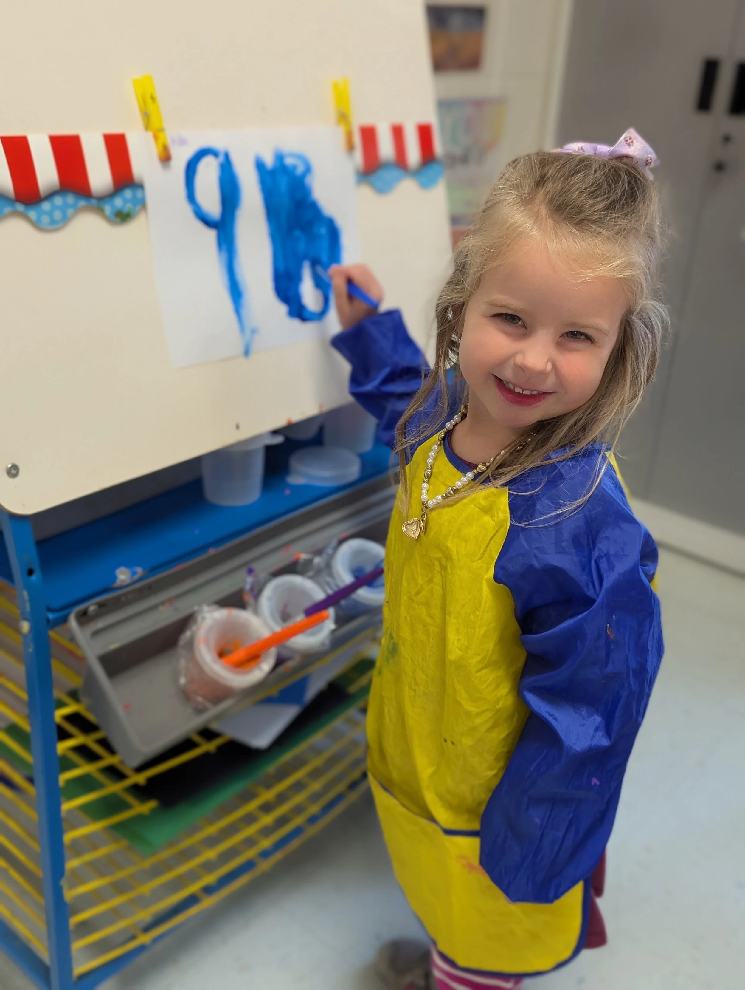Young girl painting with blue watercolors at an art station, wearing a yellow and blue smock, smiling at the camera.
