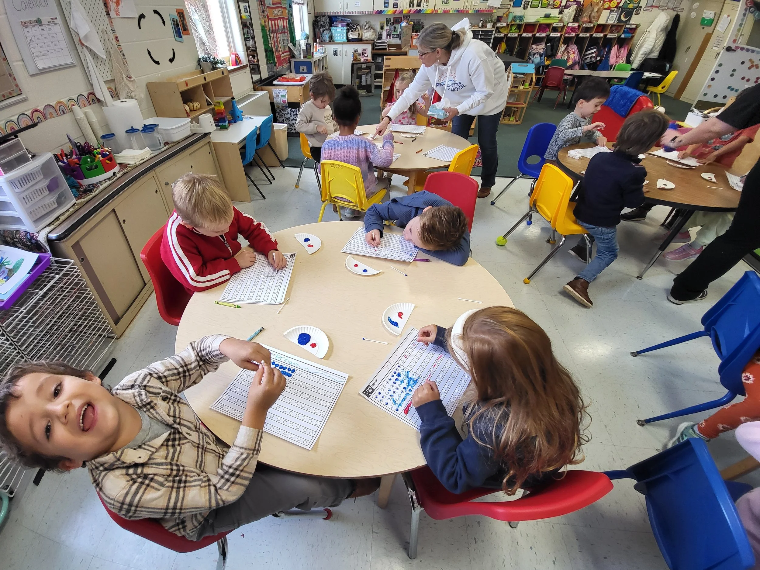 Kindergarten classroom with young children engaged in craft activities, adults assisting, colorful chairs, shelves with supplies, and educational posters.